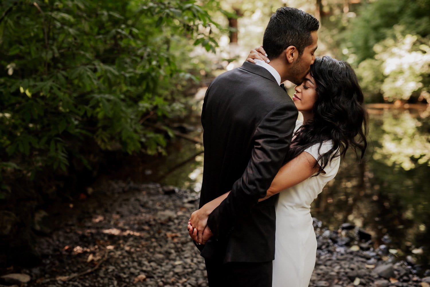 A couple in formal attire embraces by a forest stream at Leach Botanical Garden, surrounded by lush greenery, on their wedding day.