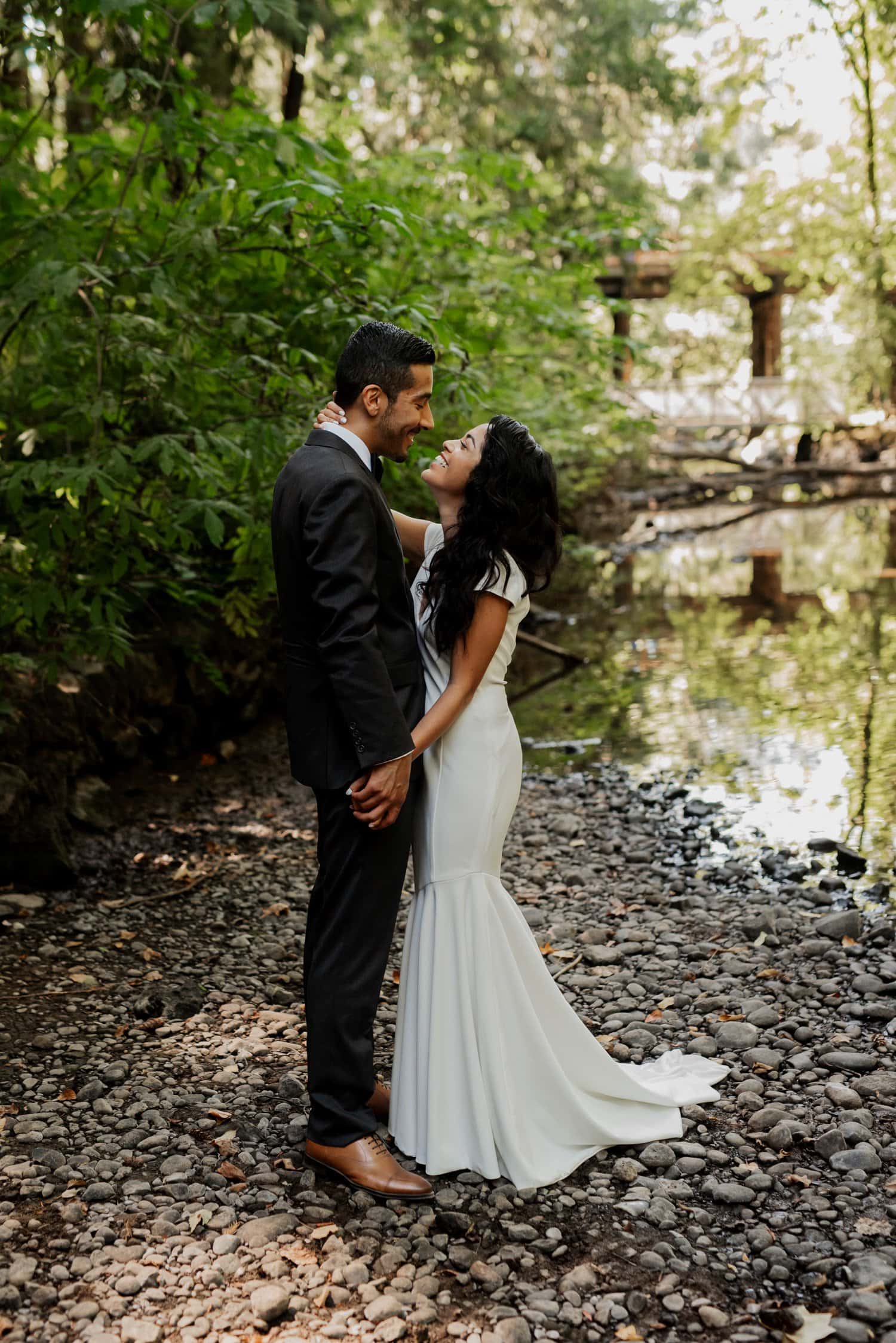 A couple embracing on a rocky forest path beside a river, with majestic trees standing tall in the background.