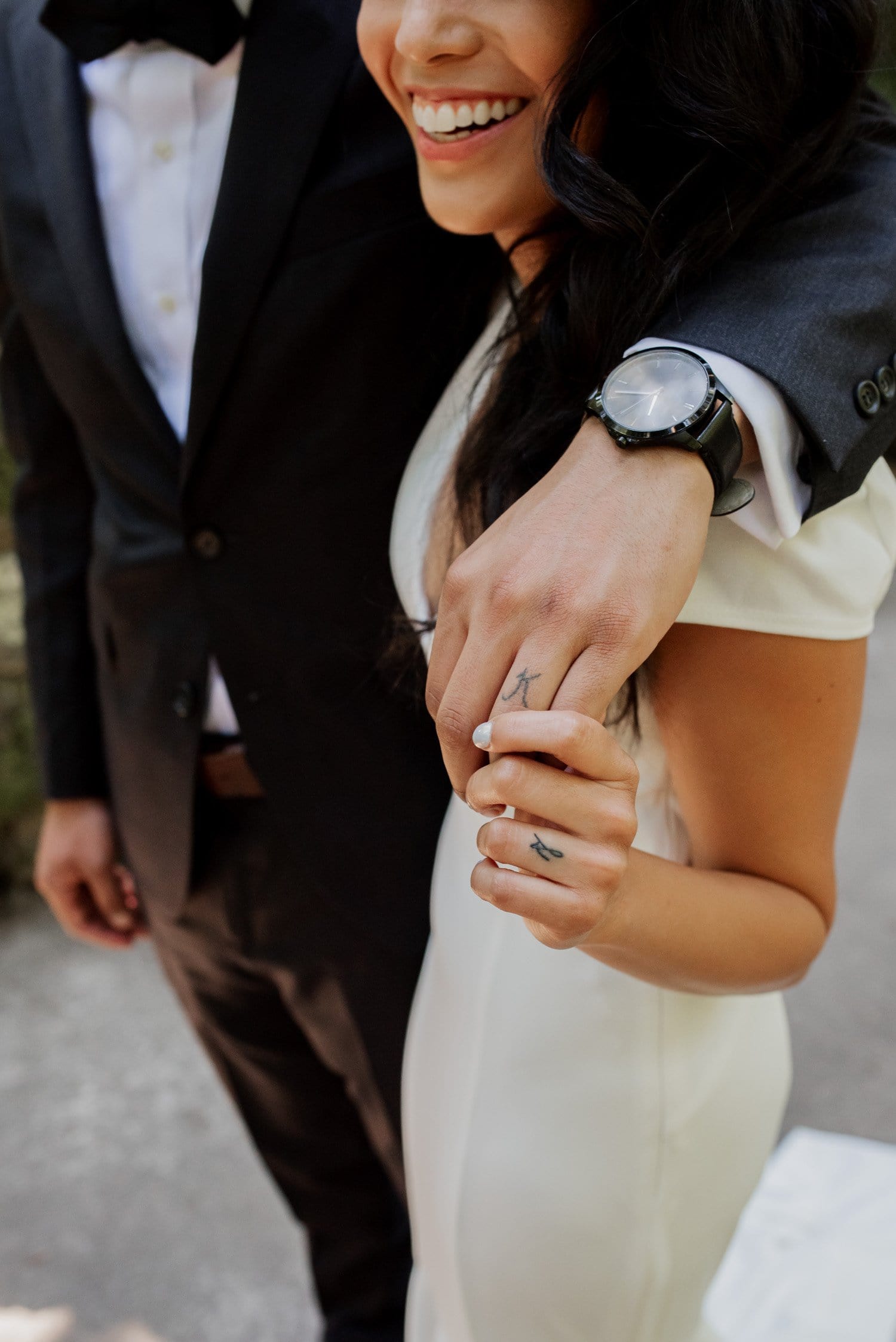 A couple stands in formal attire at Leach Botanical Garden. The man has his arm gently around the woman, who is wearing a white dress and holding his hand. Both are smiling.