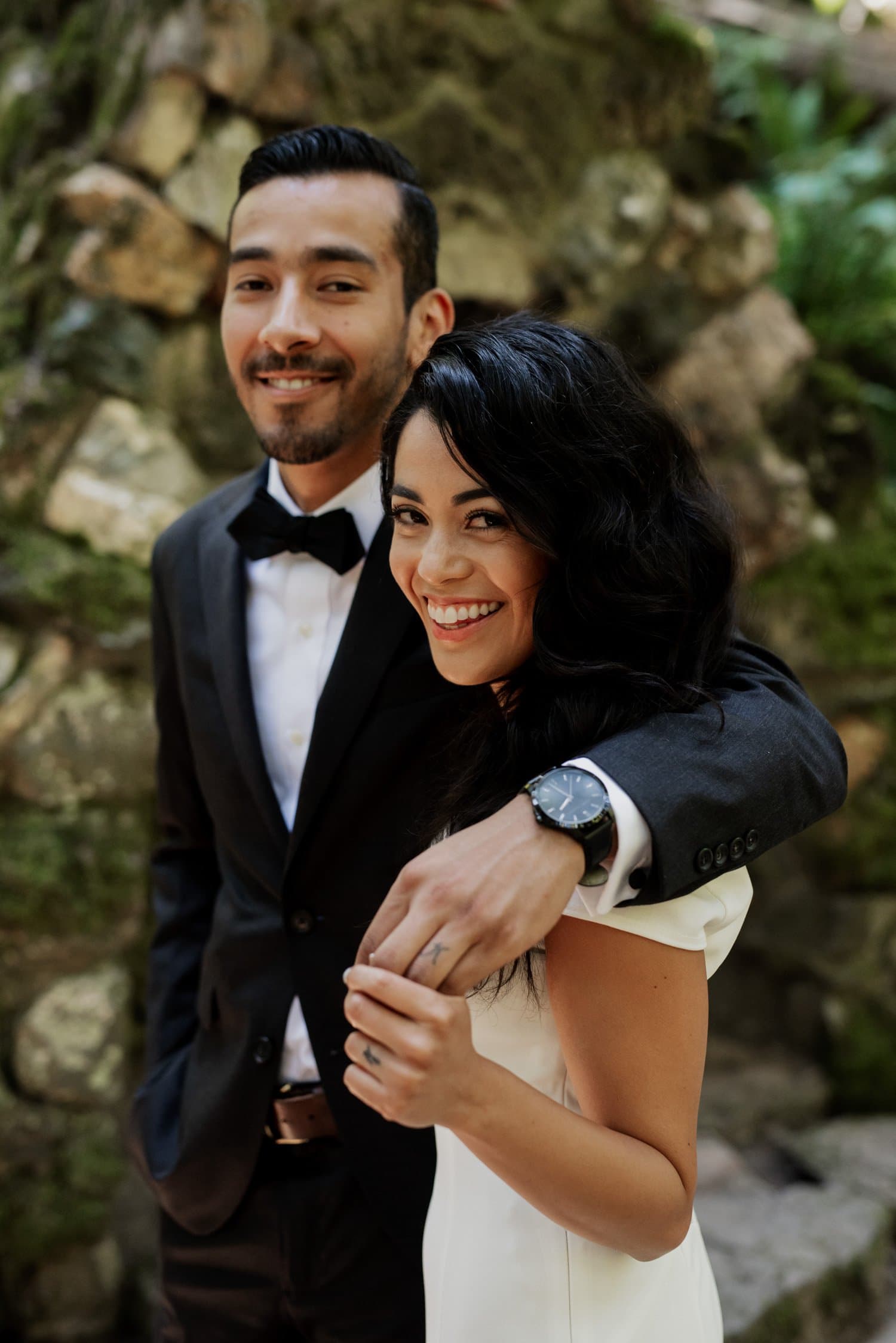 A smiling couple stands together outdoors at Leach Botanical Garden, with the man wearing a suit and bow tie and the woman in a white dress.