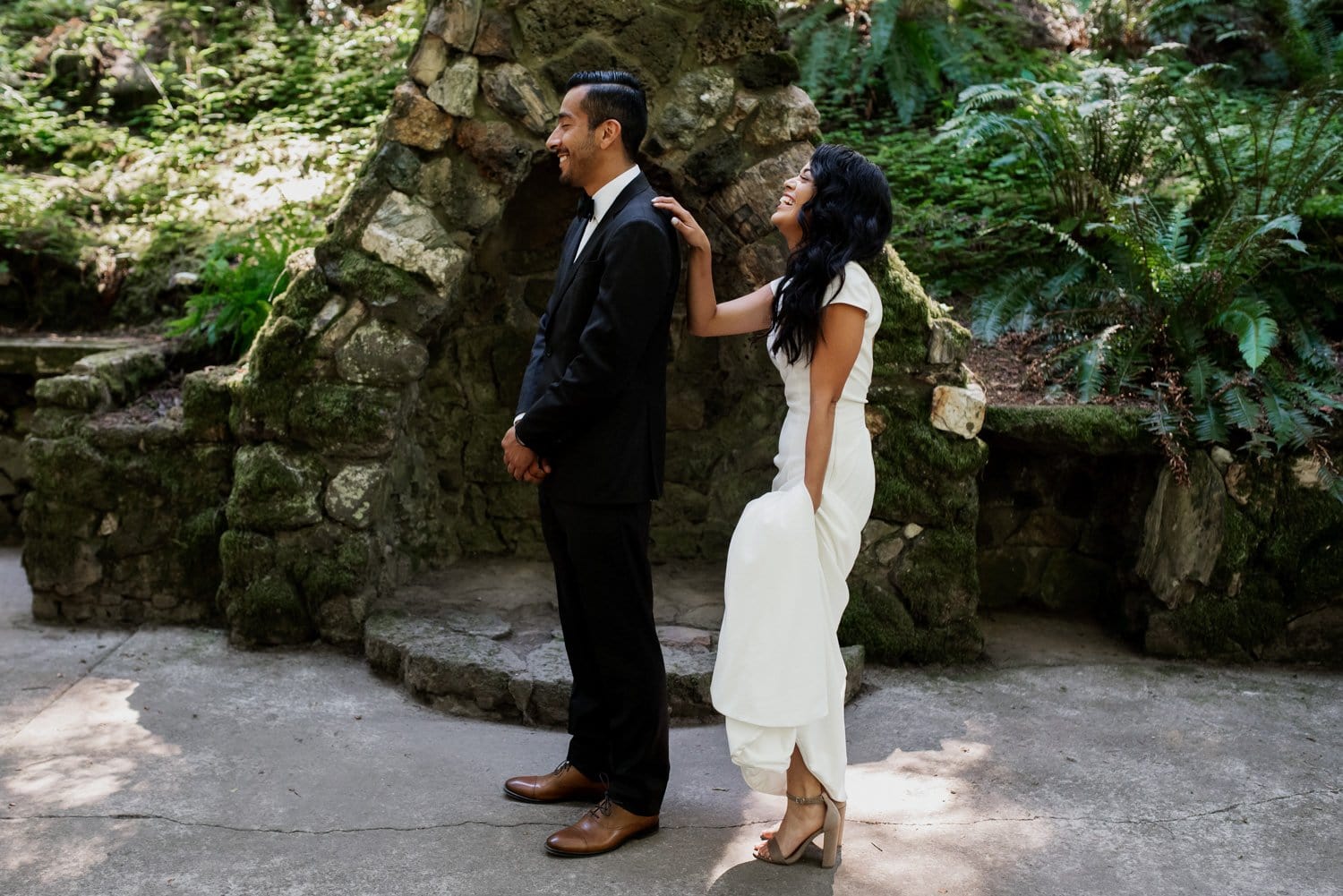 A bride in a white gown walks up behind her groom, both surrounded by lush greenery at Leach Botanical Garden.