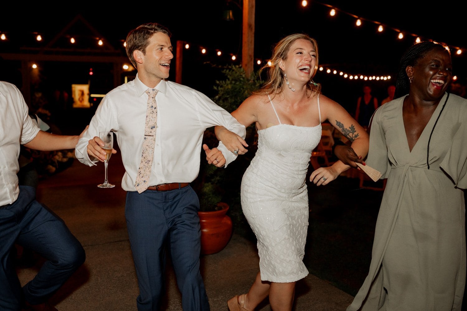 At a Cape Horn Estate wedding, people joyfully dance outside at night, holding hands. A groom in a tie dances arm-in-arm with his bride in a white dress and earrings dances beside him. String lights twinkle above, adding magic to the celebration.