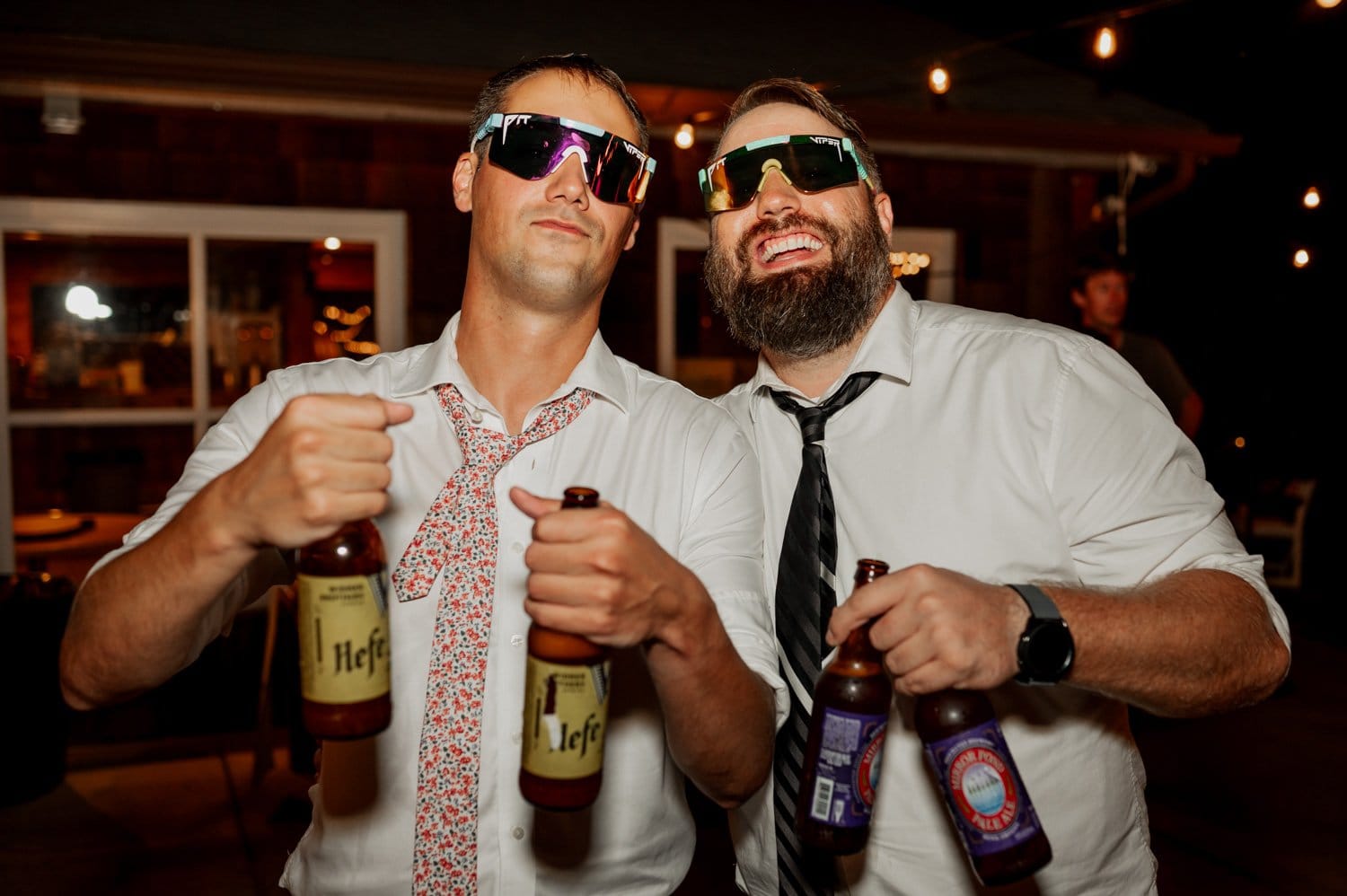 Two men in sunglasses and ties hold beer bottles, smiling for the camera amid an outdoor setting with string lights, capturing the joyous spirit of a Cape Horn Estate wedding.