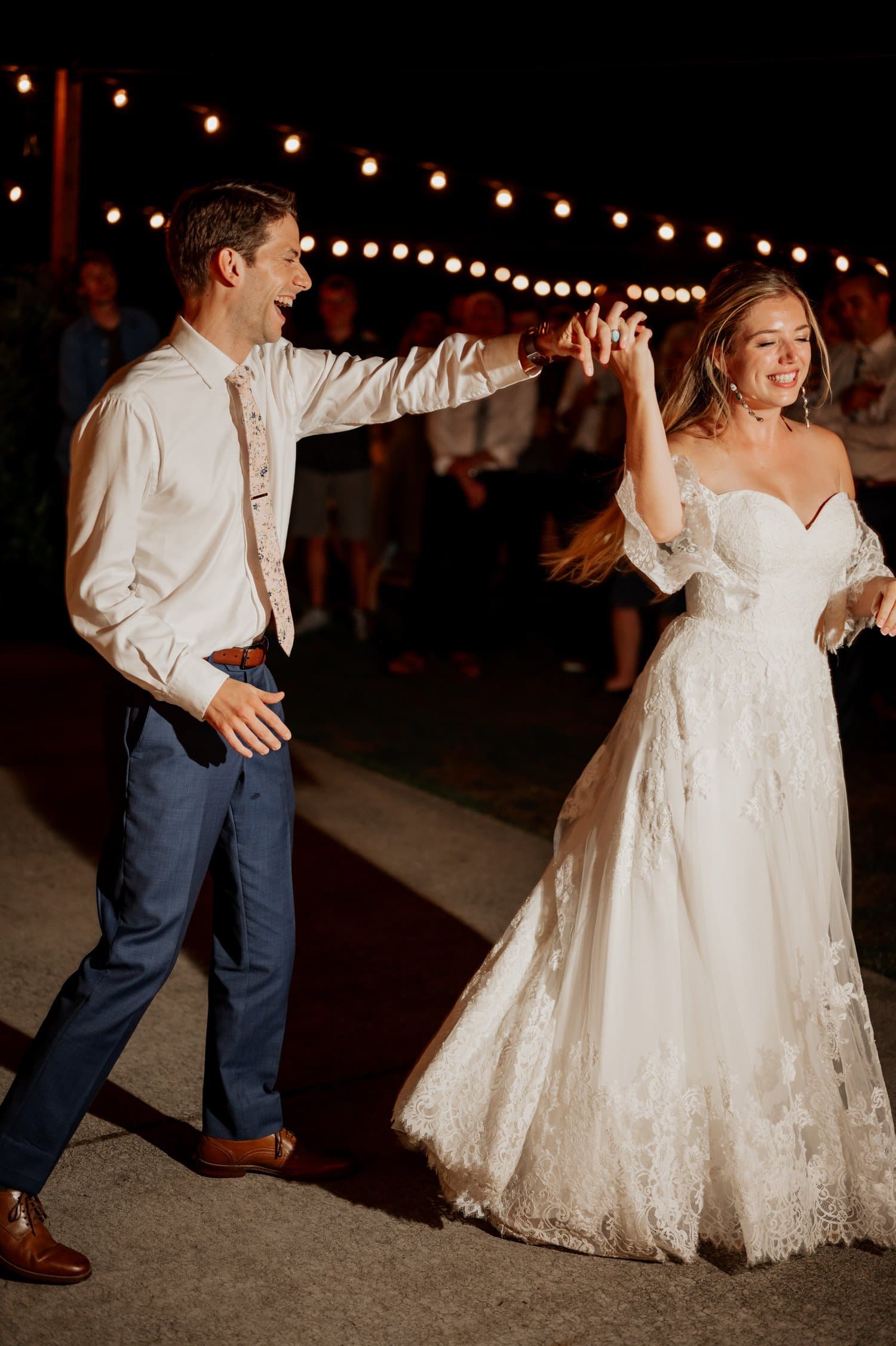 A bride in a white dress and a groom in a shirt and tie dance together under the enchanting string lights at their Cape Horn Estate wedding.