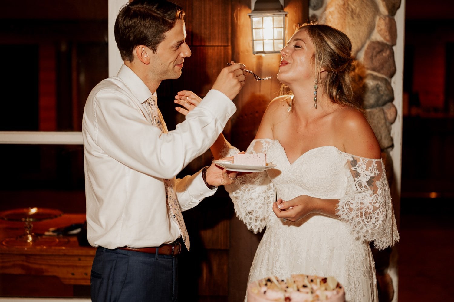 A groom in a suit feeds cake to a bride in a white lace dress at their Cape Horn Estate wedding. They stand near a stone wall and light fixture, smiling warmly at each other.