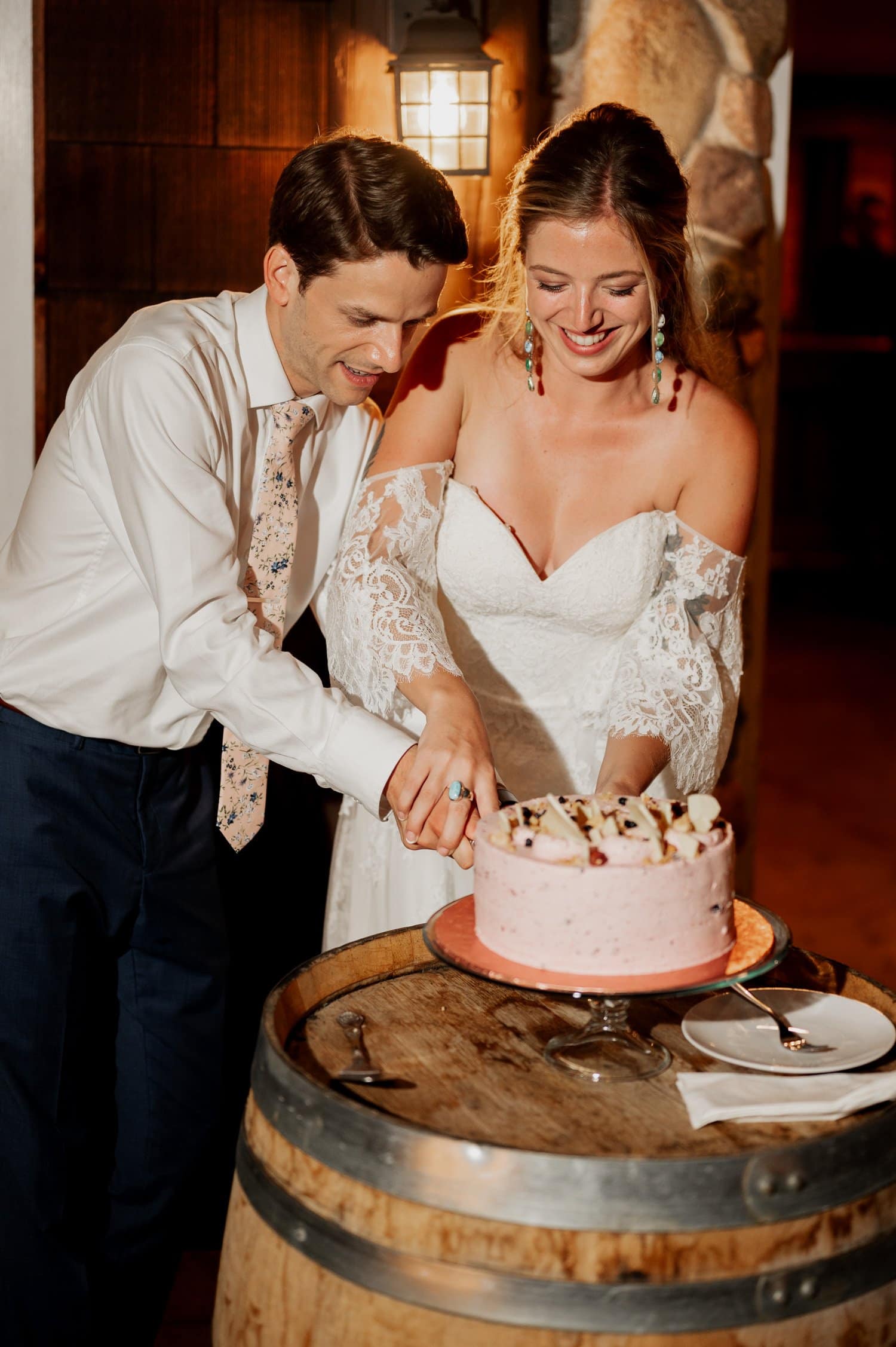 A couple in wedding attire cuts a cake together on a wooden barrel table at their Cape Horn Estate wedding, with a small lamp illuminating the background.