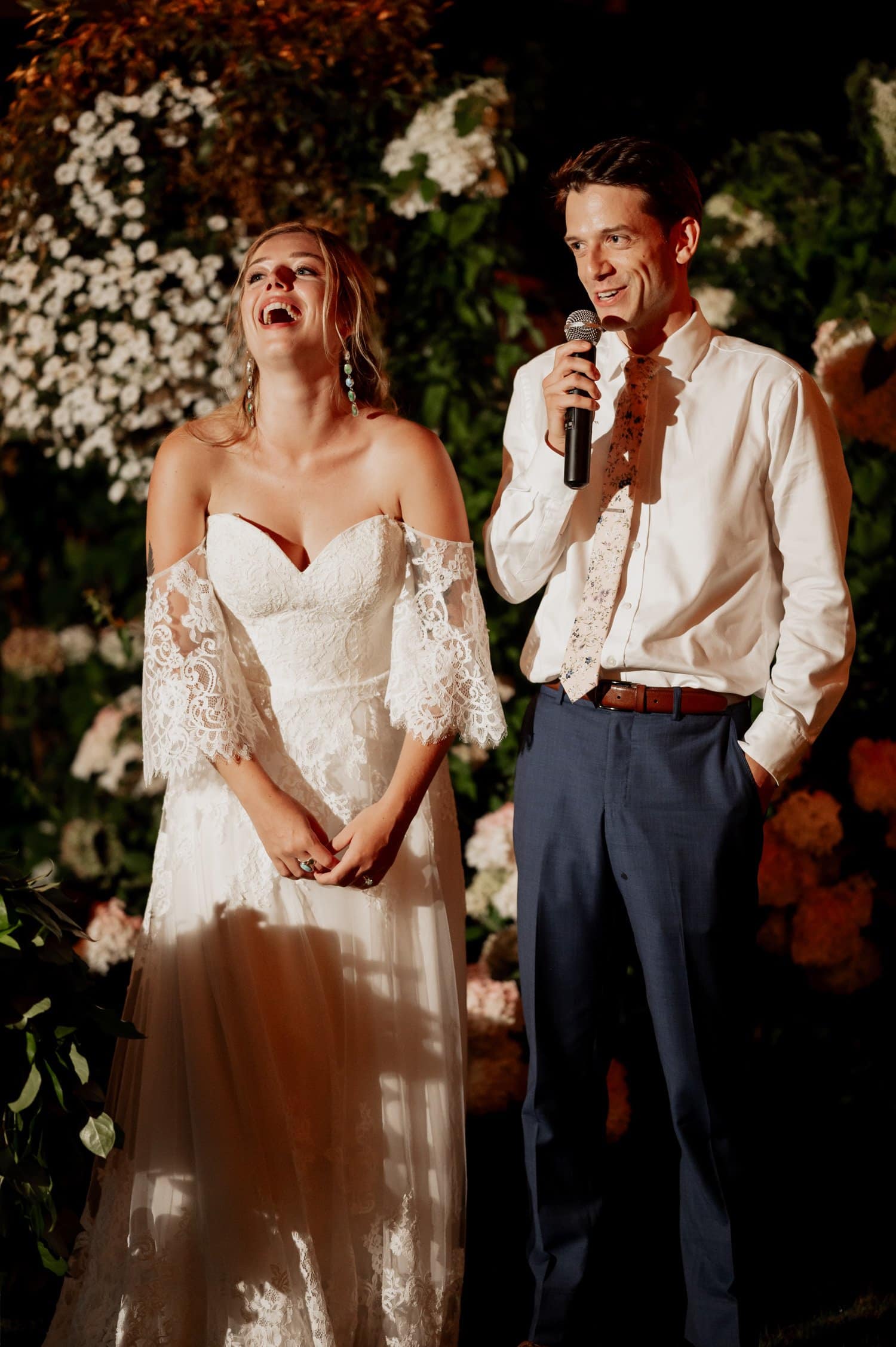 At a Cape Horn Estate wedding, a bride in a white lace dress laughs joyfully as the groom speaks into a microphone. They stand together outdoors amidst beautiful flowers and lush greenery.