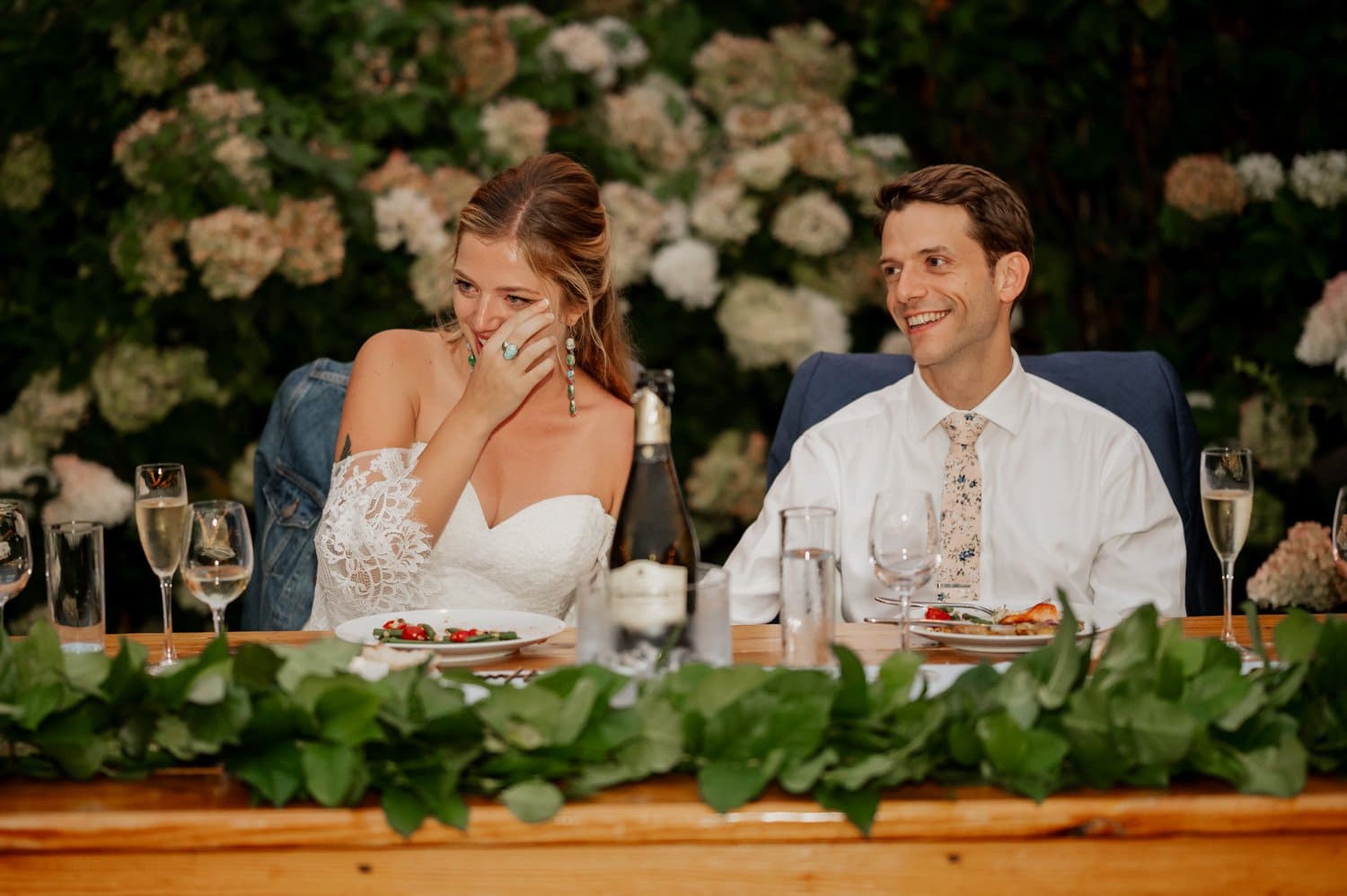 At their Cape Horn Estate wedding, the bride and groom sit at a beautifully decorated table, the bride wiping away a tear, surrounded by an array of flowers and shimmering champagne glasses.