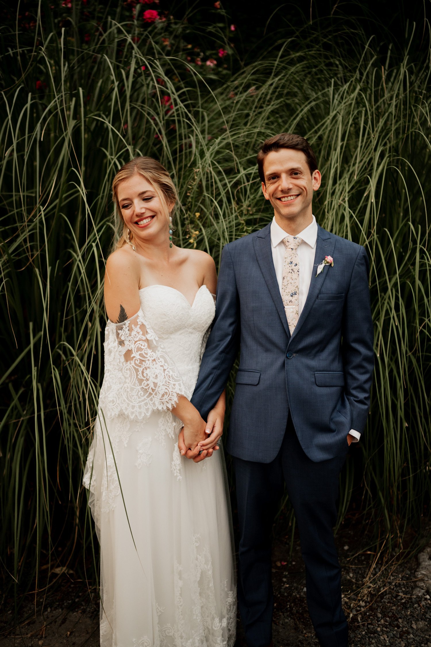 At their Cape Horn Estate wedding, the bride in a white dress and the groom in a blue suit share smiles, hand in hand, framed by tall grass.
