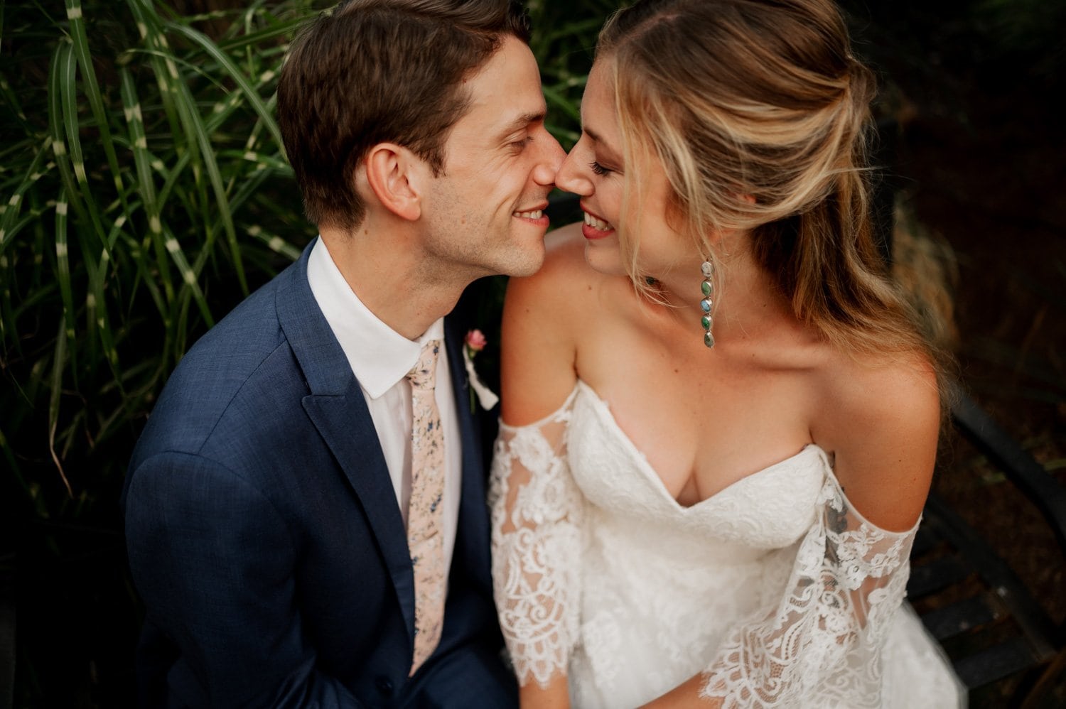 At the Cape Horn Estate wedding, a bride and groom sit closely, smiling at each other. The brides white dress with lace sleeves complements the grooms sharp blue suit, all set against a backdrop of lush greenery.