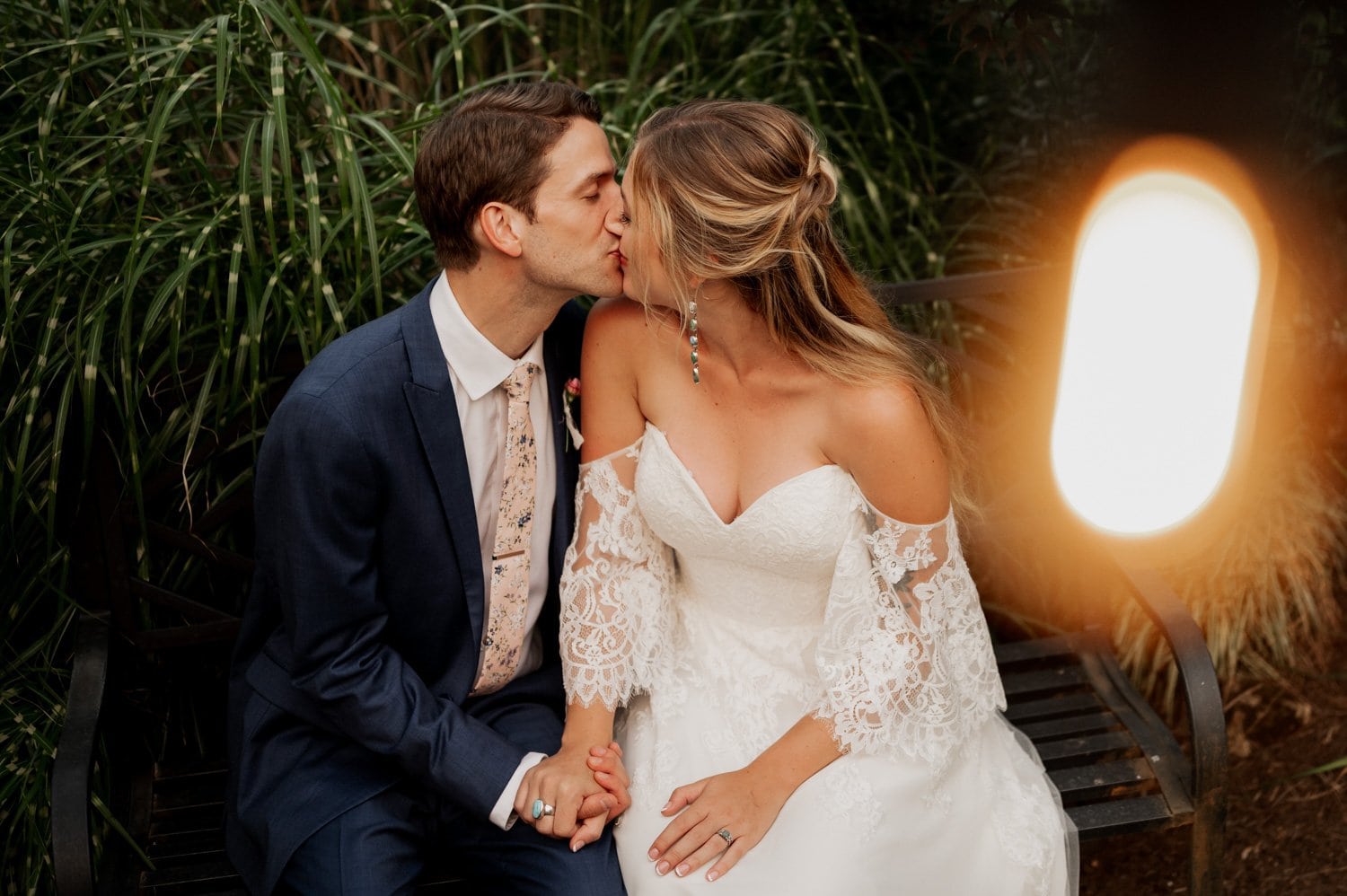 At their Cape Horn Estate wedding, the bride and groom share a kiss while seated on a bench, hands gently intertwined. She looks stunning in her white lace gown, and he is dapper in his navy suit, with decorative lighting casting a romantic glow around them.