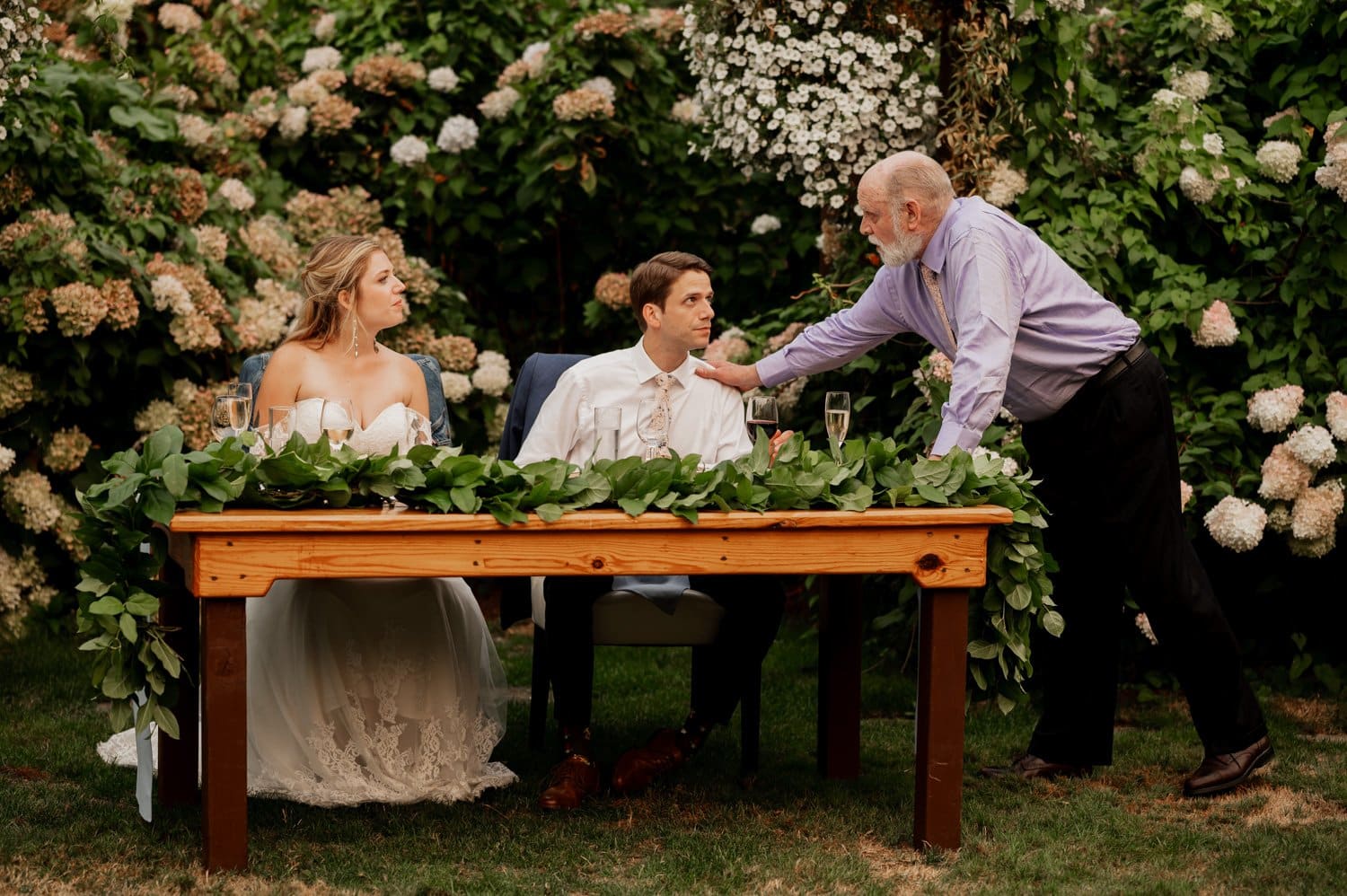 At a Cape Horn Estate wedding, a bride and groom sit at a table adorned with greenery. An older man leans in to chat, enveloped by lush foliage and white flowers, creating an enchanting backdrop.