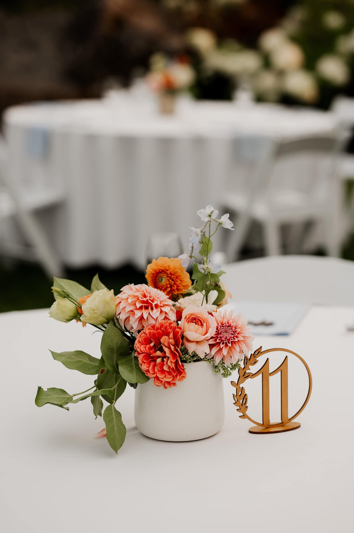 A floral centerpiece with orange and pink blooms adorns the Cape Horn Estate wedding table, held elegantly in a white vase beside a metallic number 11 table marker on a pristine white tablecloth.
