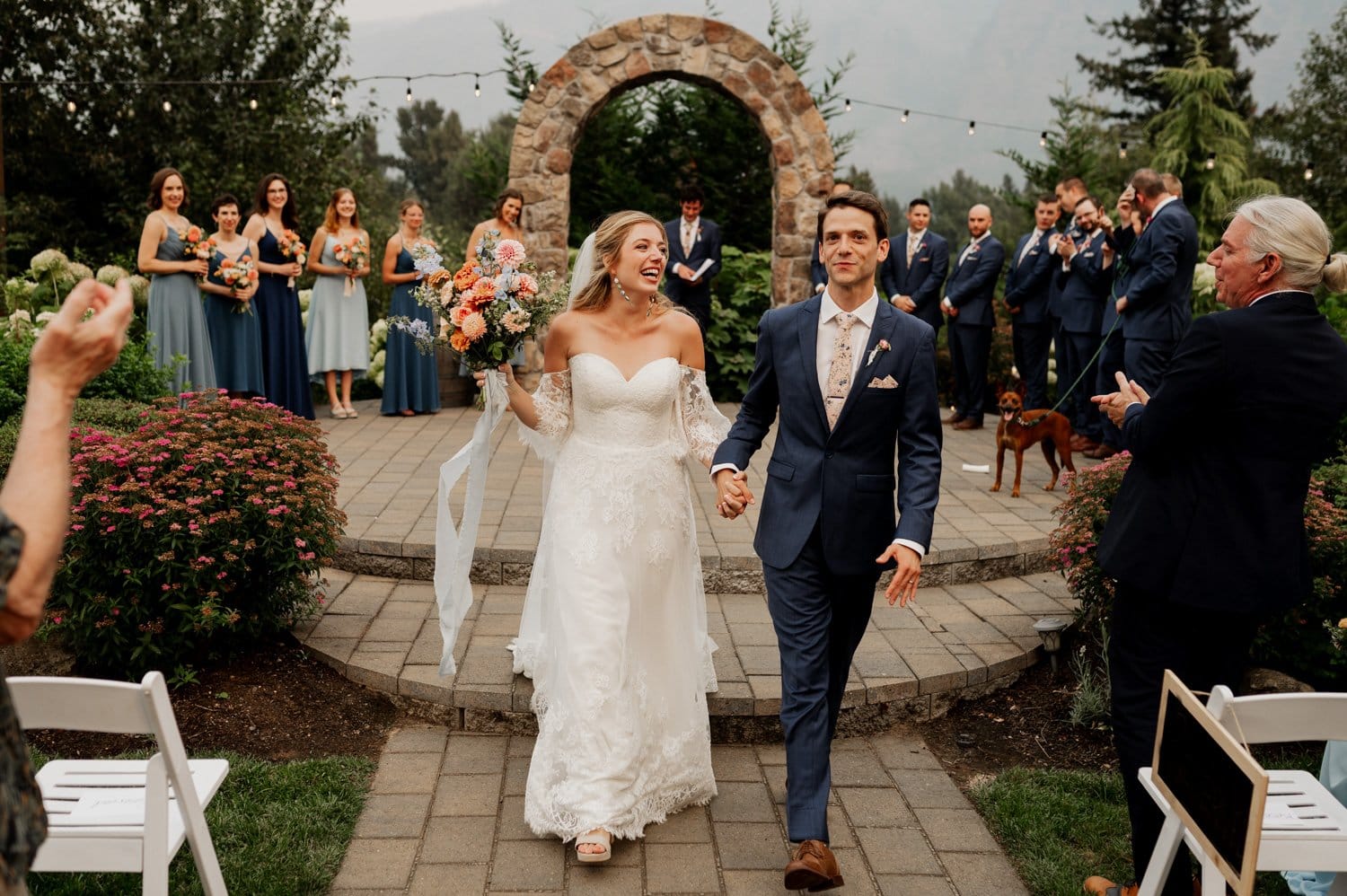 The bride and groom walk joyfully down the aisle at their Cape Horn Estate wedding, under a picturesque stone archway, as the wedding party and guests applaud.