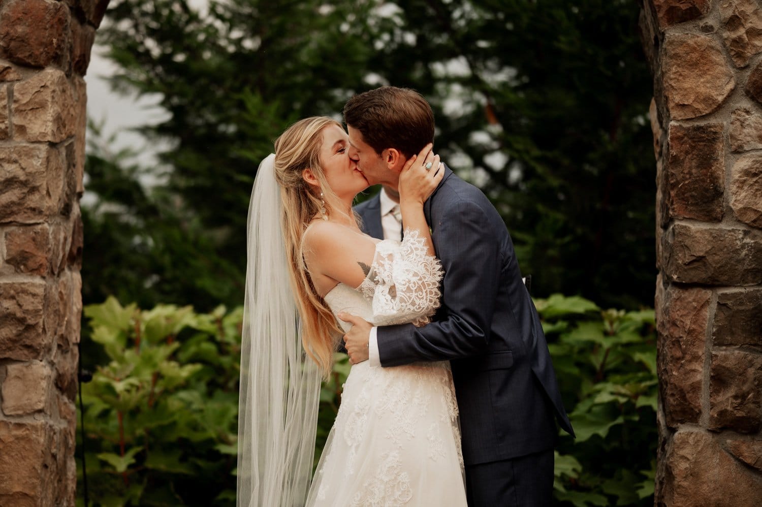 A couple in wedding attire shares a kiss outdoors at Cape Horn Estate, framed by stone arches and lush greenery.