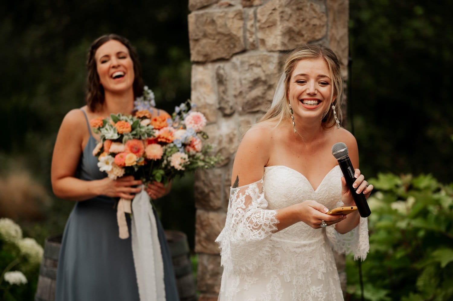 At a Cape Horn Estate wedding, a bride in a white lace dress holds a microphone and smiles. Beside her, a bridesmaid in a blue dress clutches a colorful bouquet, laughing gleefully. They stand outside near the rustic charm of an old stone pillar.