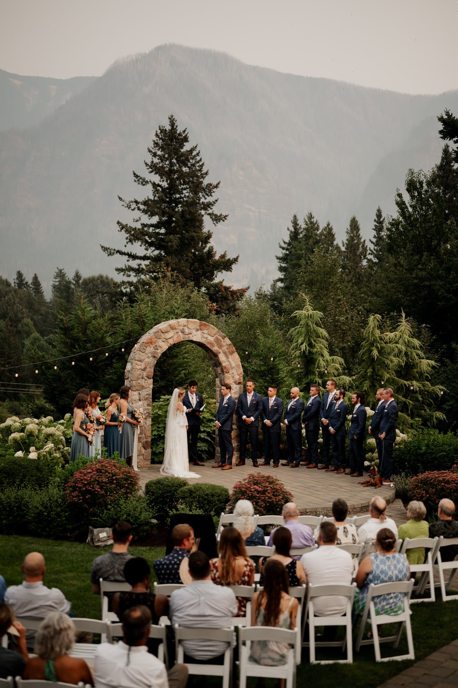 At a Cape Horn Estate wedding, a couple stands at an altar beneath a stone arch. Bridesmaids and groomsmen are perfectly lined up on either side, while guests sit attentively in the foreground, framed by the lush forested mountains stretching into the horizon.