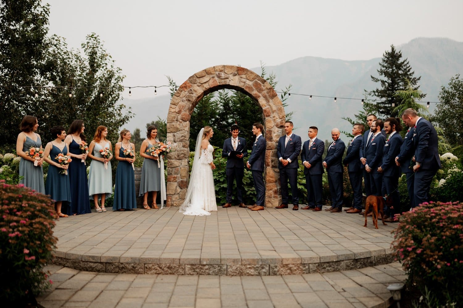 A picturesque Cape Horn Estate wedding unfolds as the bride and groom stand under a stone arch. Bridesmaids and groomsmen, elegantly clad in blue, line each side while a charming dog sits on the right, adding a delightful touch to this outdoor celebration.