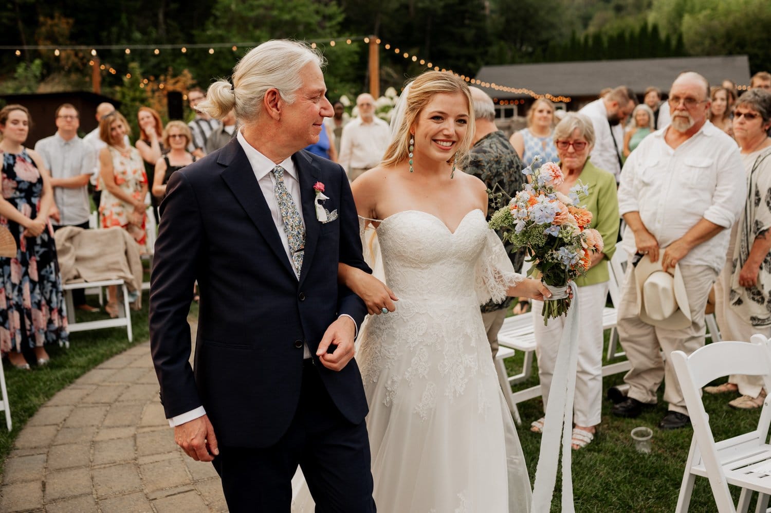 At a Cape Horn Estate wedding, the bride in a white gown holds a bouquet as she walks arm-in-arm with her father in a suit down the outdoor aisle, surrounded by seated and standing guests.