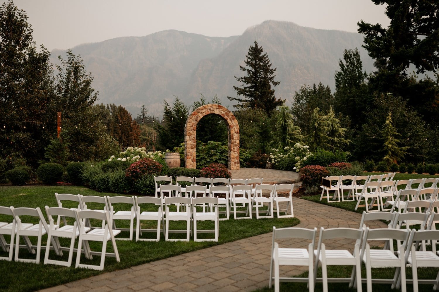 Outdoor wedding setup at Cape Horn Estate with white chairs arranged on a lawn facing a brick archway surrounded by lush greenery, with majestic mountains visible in the background.