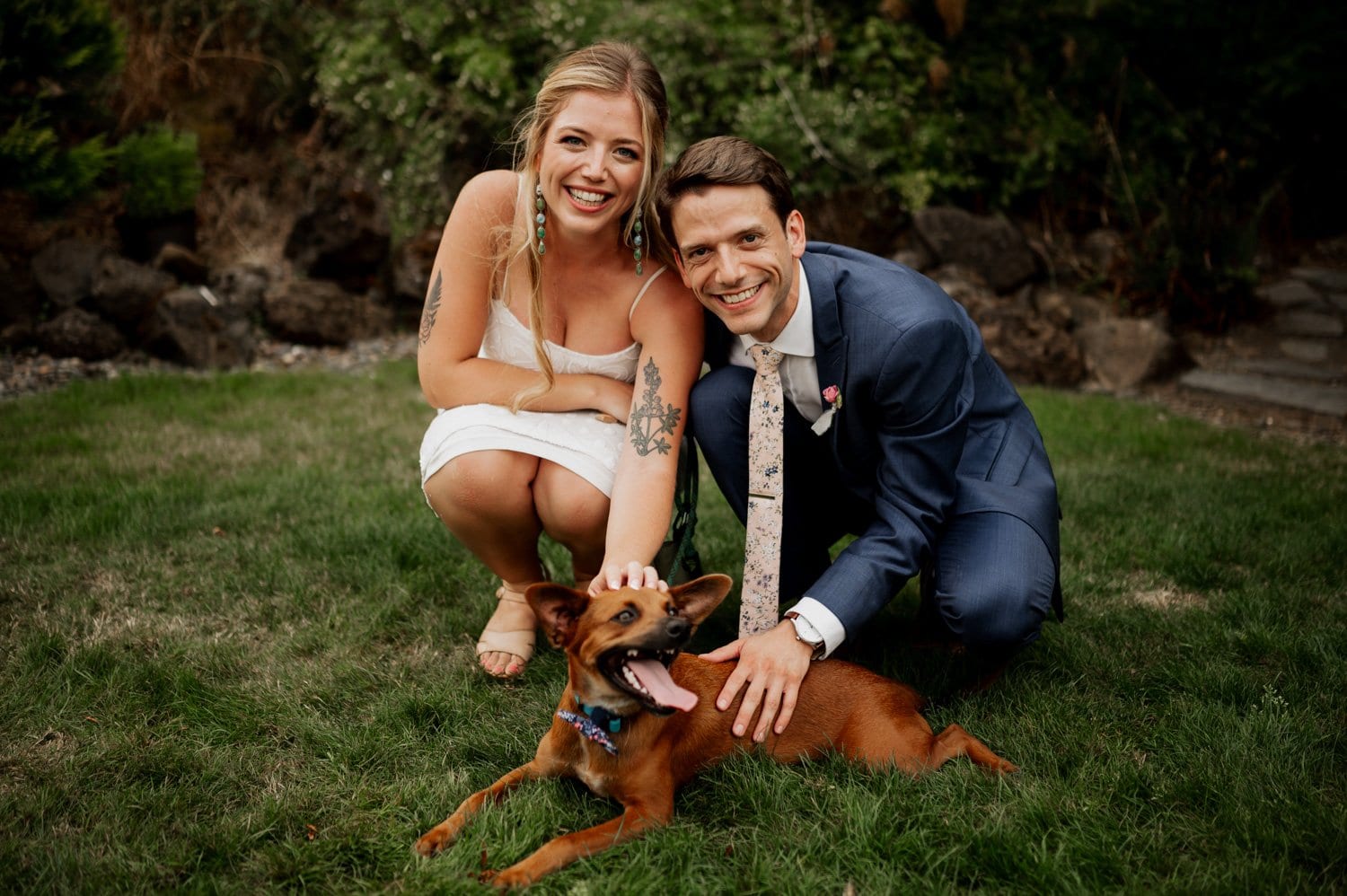 A bride and groom in formal attire kneels on the grass at their Cape Horn Estate wedding, petting a small brown dog lying in front of them, with trees and rocks adding to the picturesque backdrop.