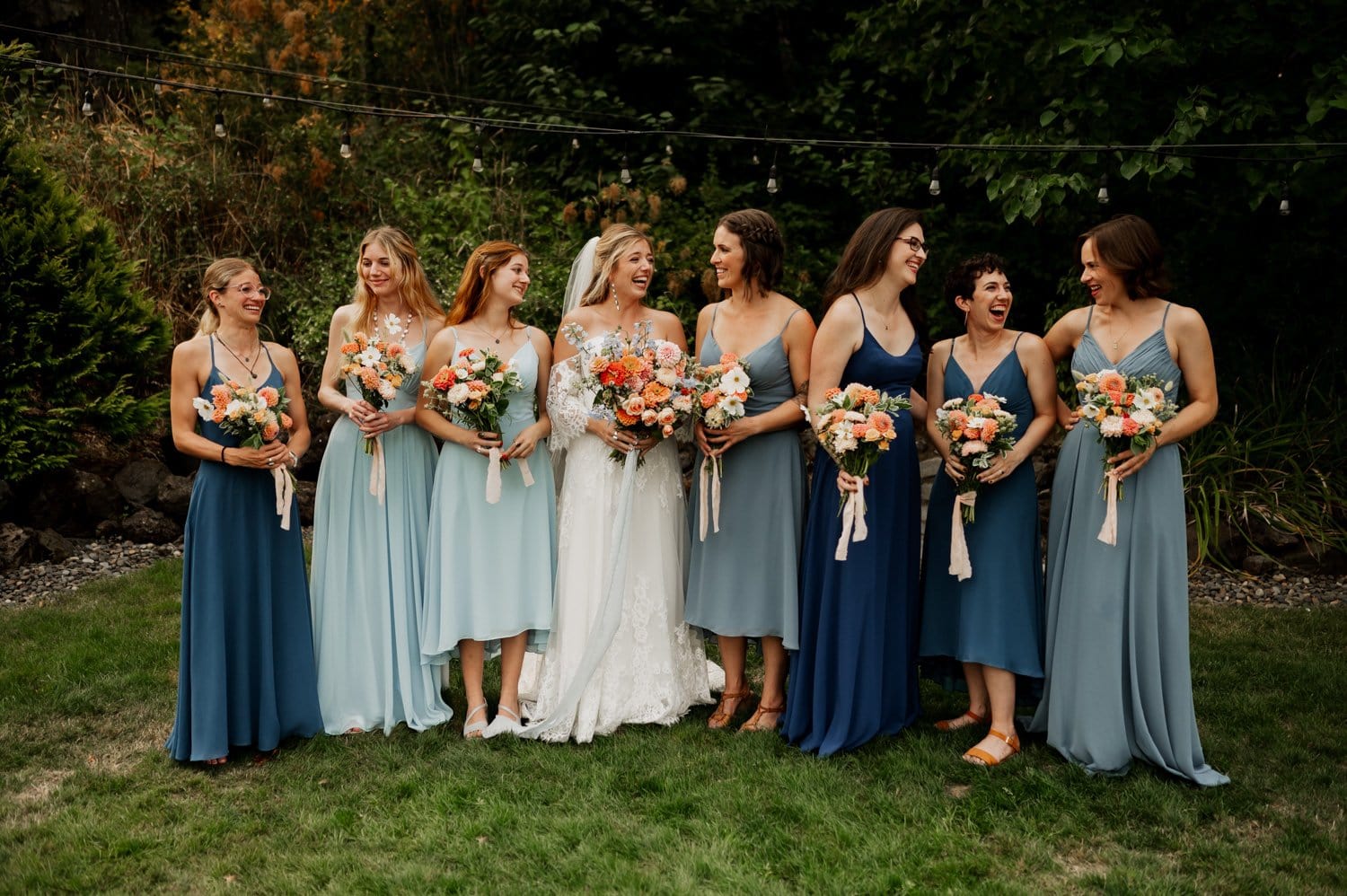 A bride in a white gown stands with seven bridesmaids in blue and teal dresses, all holding bouquets. They are outdoors on the lush grounds of Cape Horn Estate, with towering trees providing a picturesque backdrop.