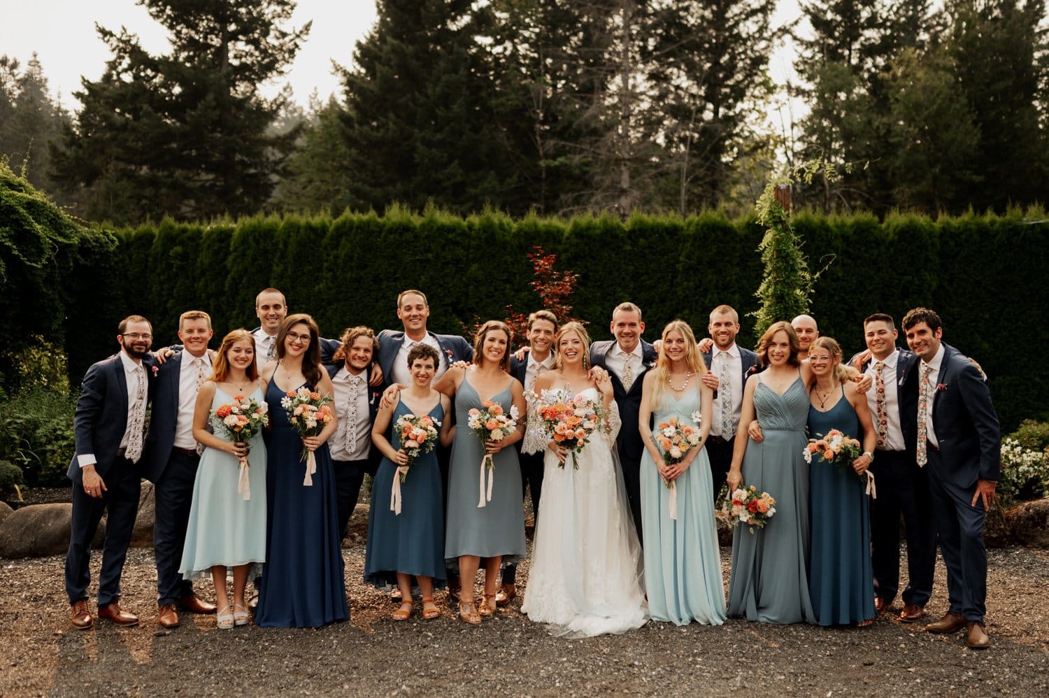 The wedding party stands outdoors at Cape Horn Estate, with bridesmaids in various shades of blue dresses and groomsmen in navy suits. The bride and groom are centered, holding bouquets, with a backdrop of lush trees completing the picturesque scene.