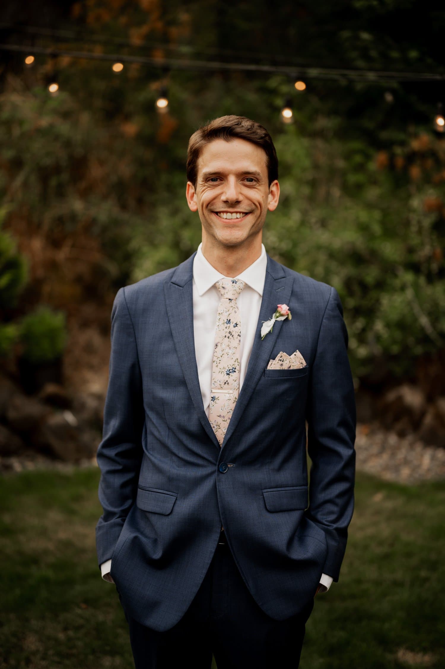 A groom in a blue suit and floral tie stands outdoors at Cape Horn Estate, smiling with hands in pockets. The enchanting backdrop features lush greenery and twinkling string lights, perfect for the magical wedding ambiance.