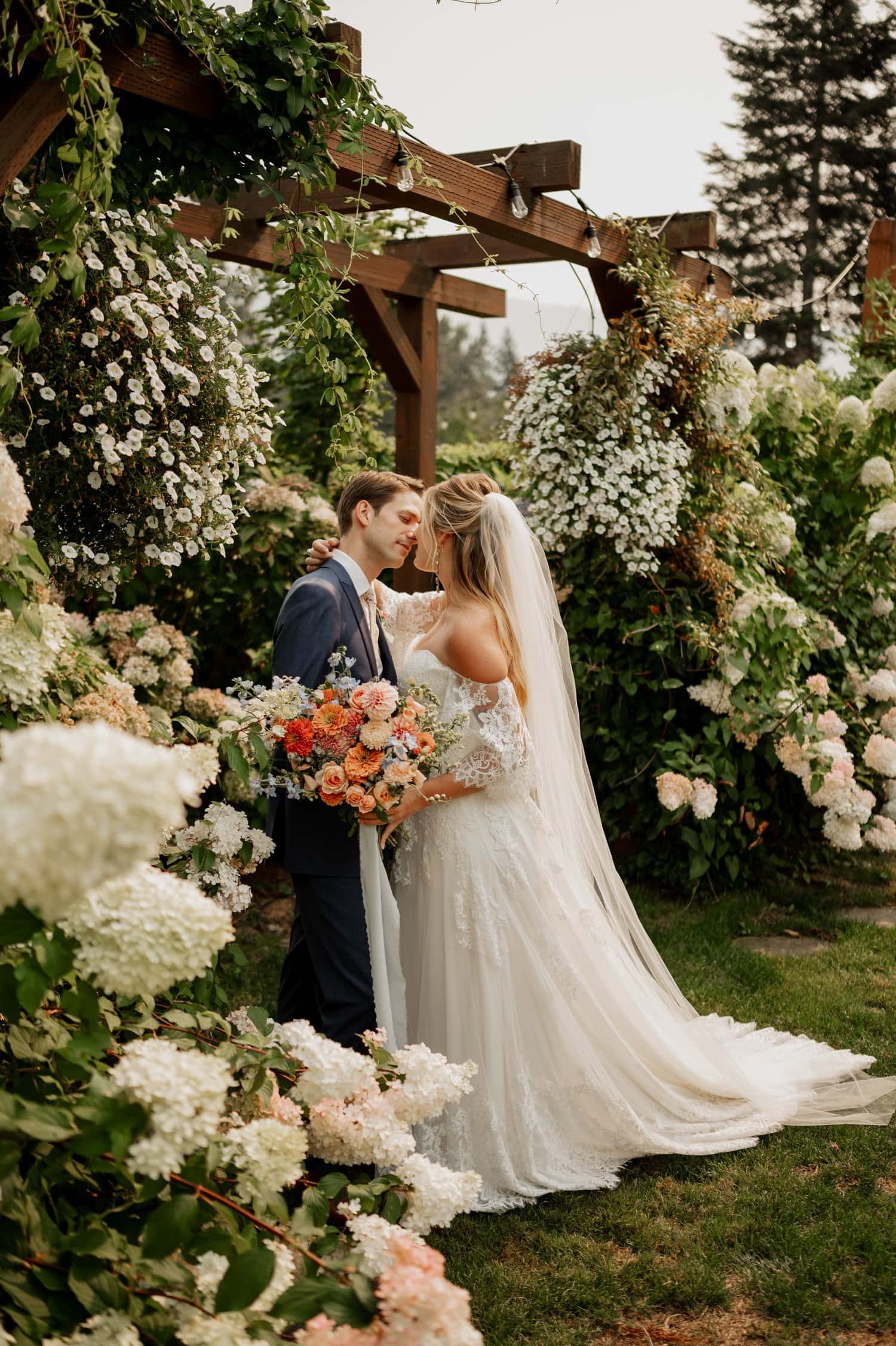 A bride and groom stand together in a lush garden at Cape Horn Estate, surrounded by white and pink flowers under a wooden pergola. The bride holds a bouquet, and both are dressed in formal wedding attire.