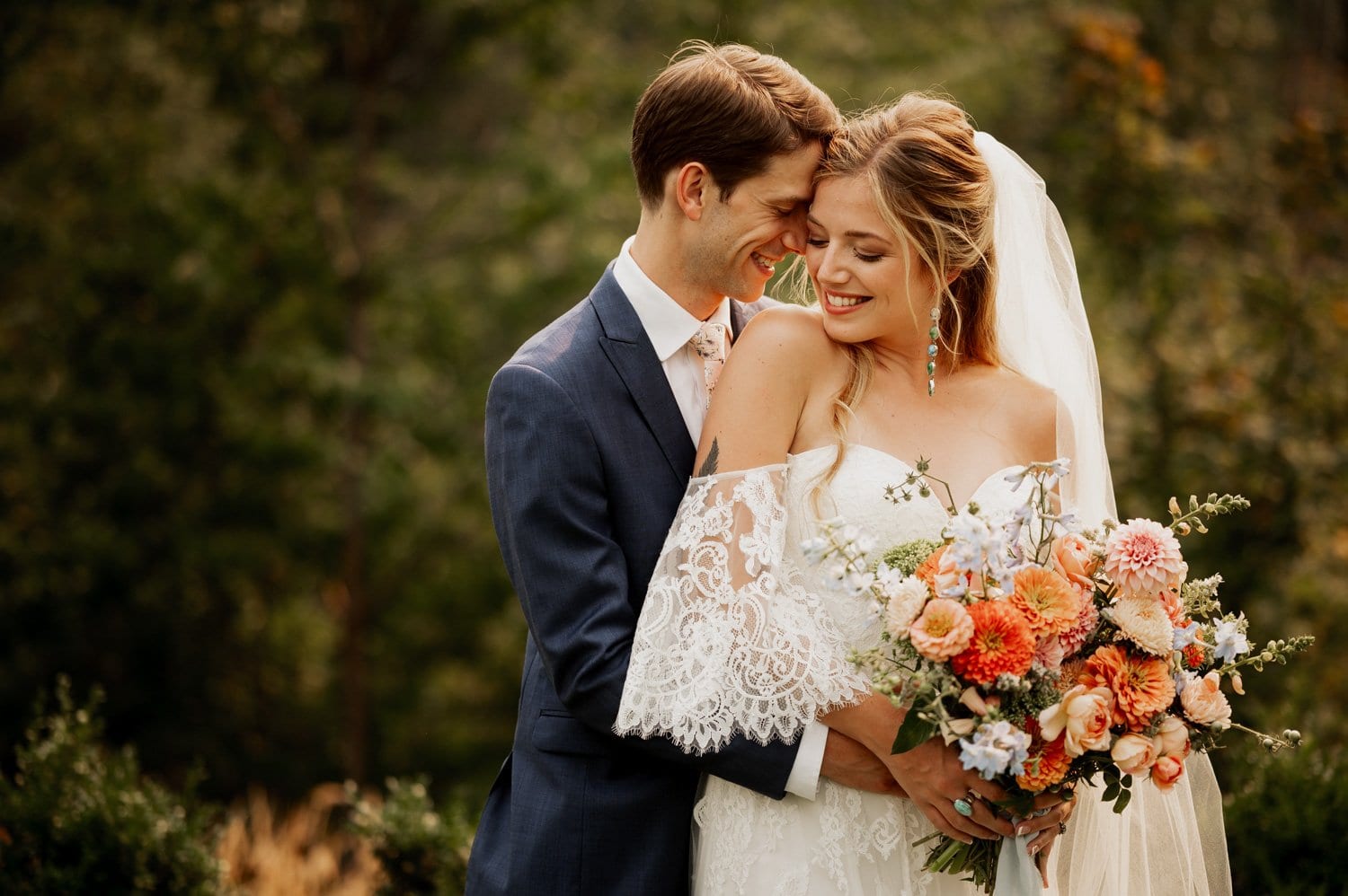 A bride holding a bouquet is embraced by a groom in a blue suit, both smiling, with the enchanting forest backdrop of Cape Horn Estate painting a perfect picture of their wedding day.