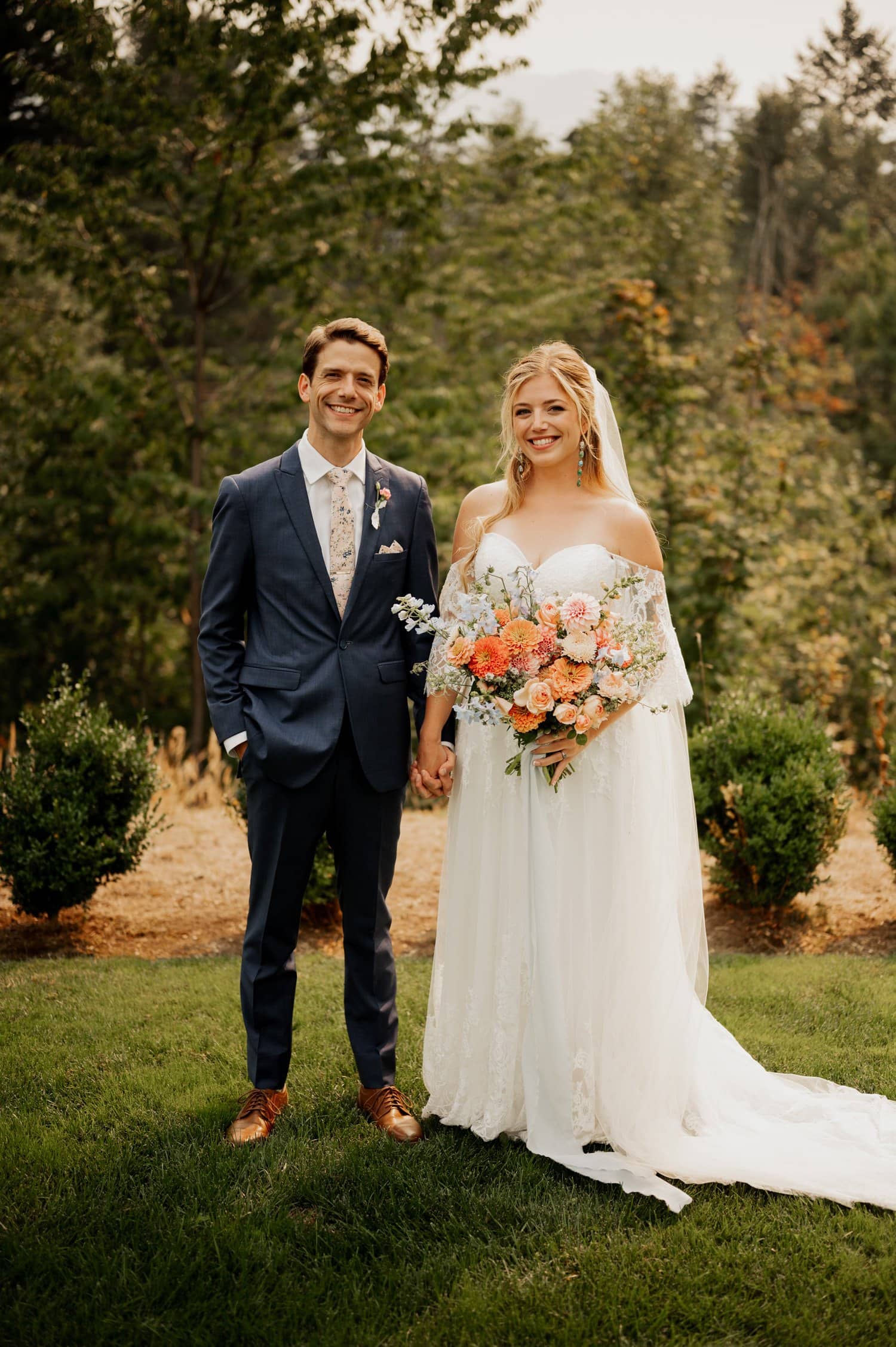 At their Cape Horn Estate wedding, the bride in a white gown holding a bouquet stands beside the groom in a blue suit. Both are smiling, surrounded by the enchanting backdrop of trees.