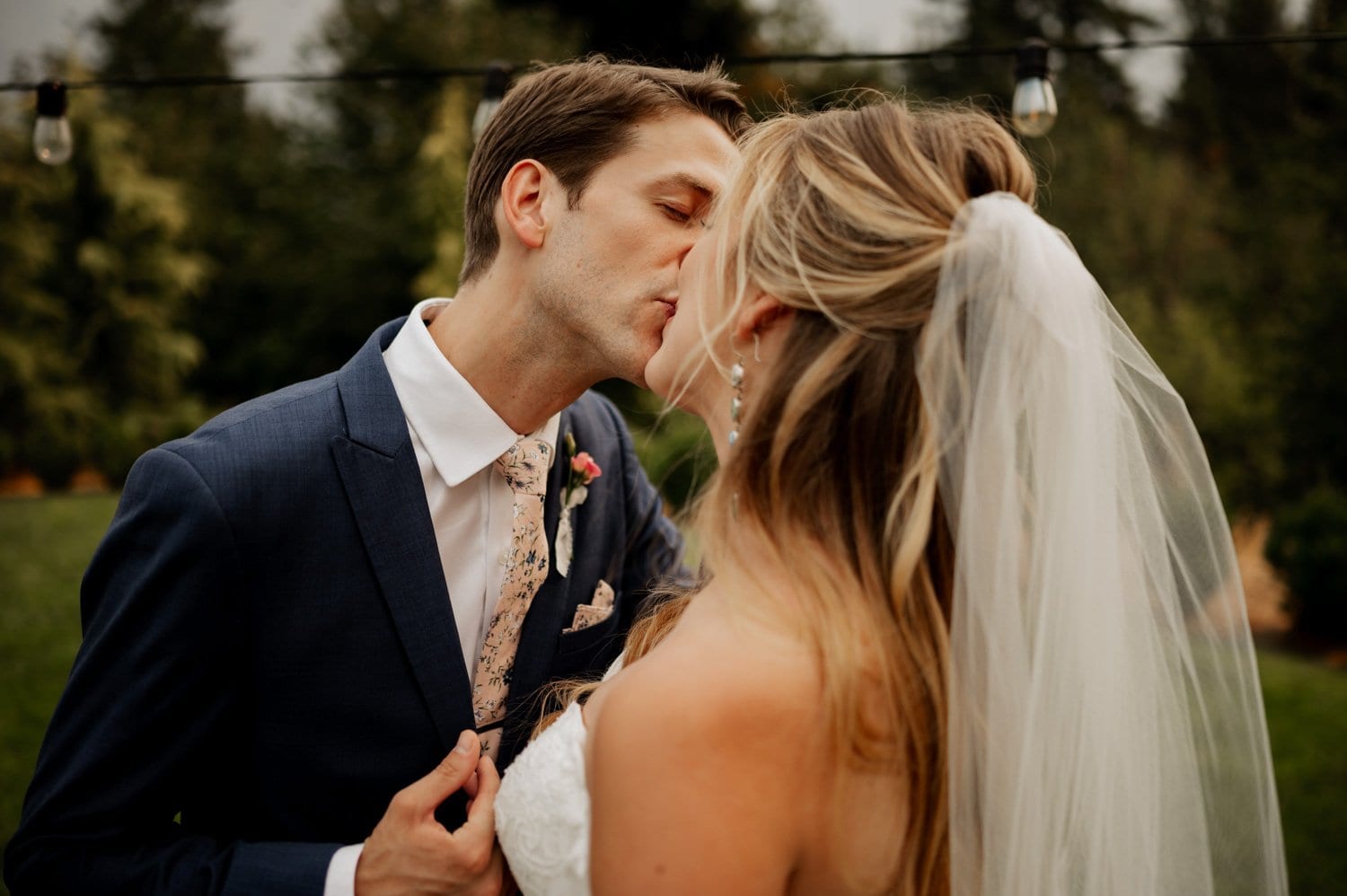 At their Cape Horn Estate wedding, a couple shares a tender kiss outdoors. The man dons a navy suit with a floral tie, while the woman wears an elegant white dress and veil. Soft string lights twinkle above them, capturing the enchanting moment.