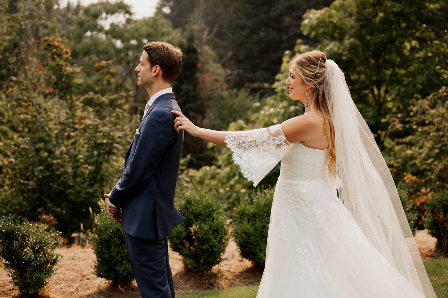 At a Cape Horn Estate wedding, a bride in a white dress and veil gently taps the shoulder of her groom in a blue suit. Surrounded by lush trees and bushes, their outdoor moment is one of pure enchantment.