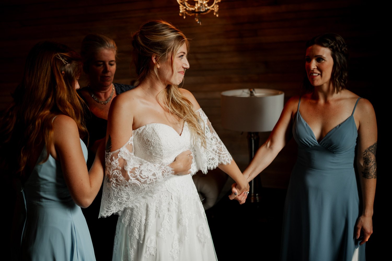 A bride in a lace gown, at a Cape Horn Estate wedding, is surrounded by three women, all wearing blue dresses, in a warmly lit room.