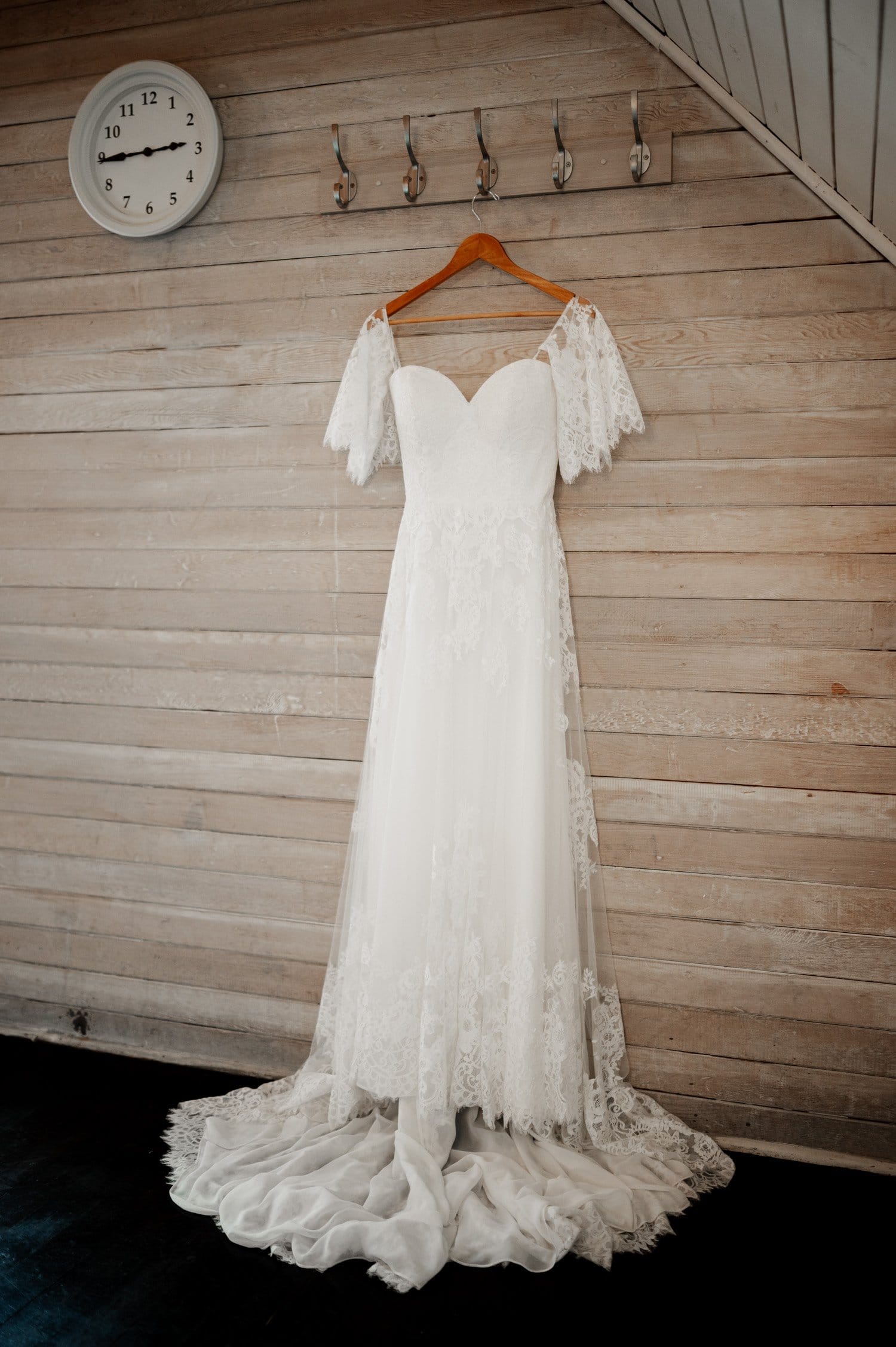 White lace wedding dress with a sweetheart neckline and short sleeves hangs elegantly on a wooden wall at the picturesque Cape Horn Estate. A round clock above, marking 11:12, adds to the serene anticipation of the day.
