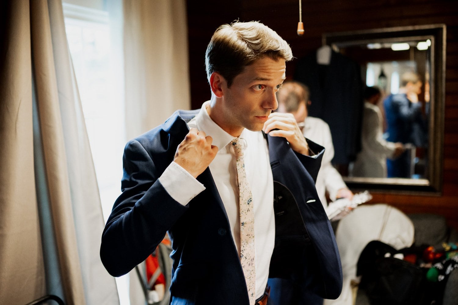 A groom adjusts his suit jacket in a room with mirrored walls and curtains, the elegant setting hinting at the grandeur of a Cape Horn Estate wedding. Another person is visible in the reflection, adding to the scenes anticipation.