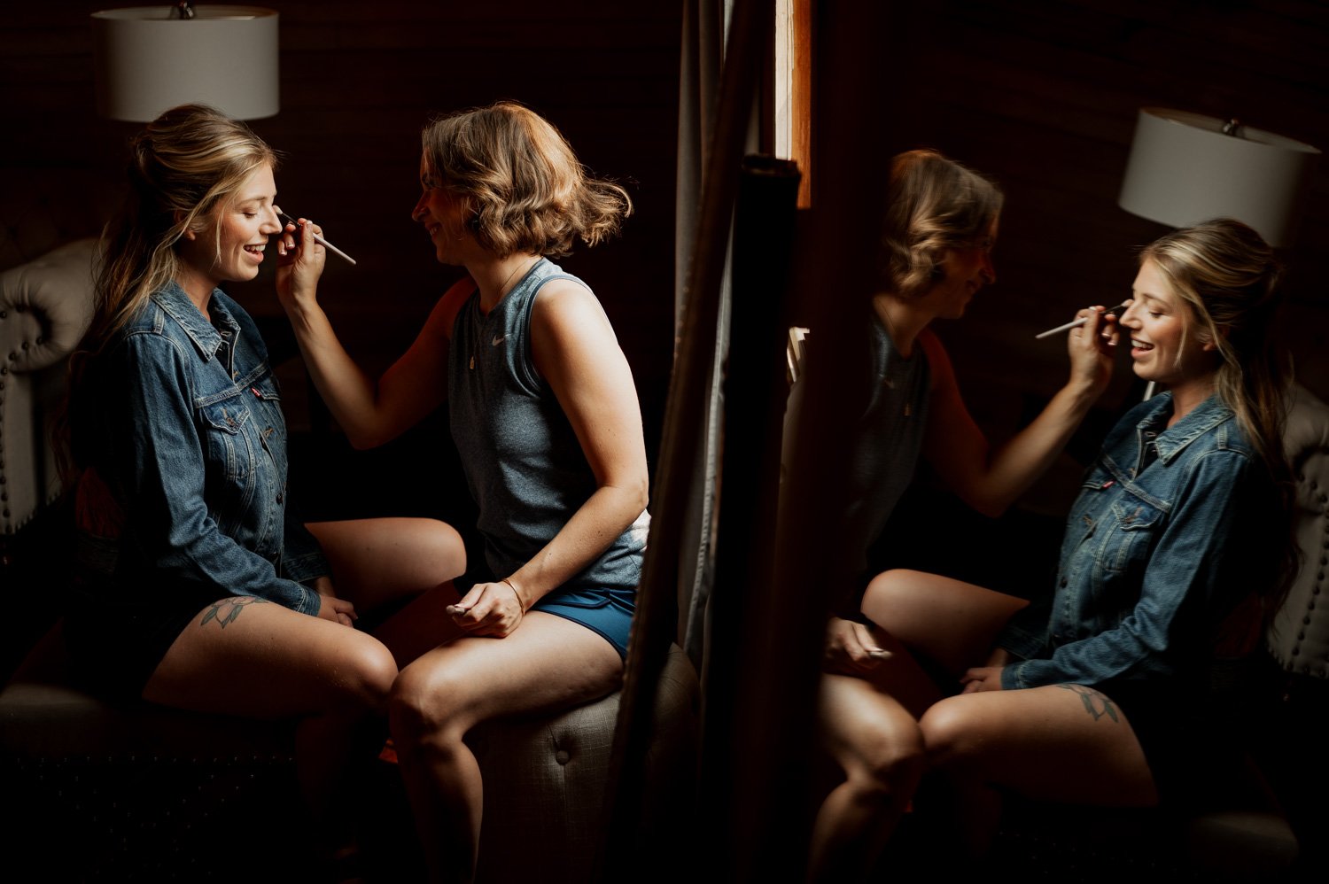 Two women are seated, one applying makeup to the other as they prepare for the Cape Horn Estate wedding. They are in a dimly lit room with a mirror reflecting the scene. One wears a denim jacket; the other a sleeveless top.