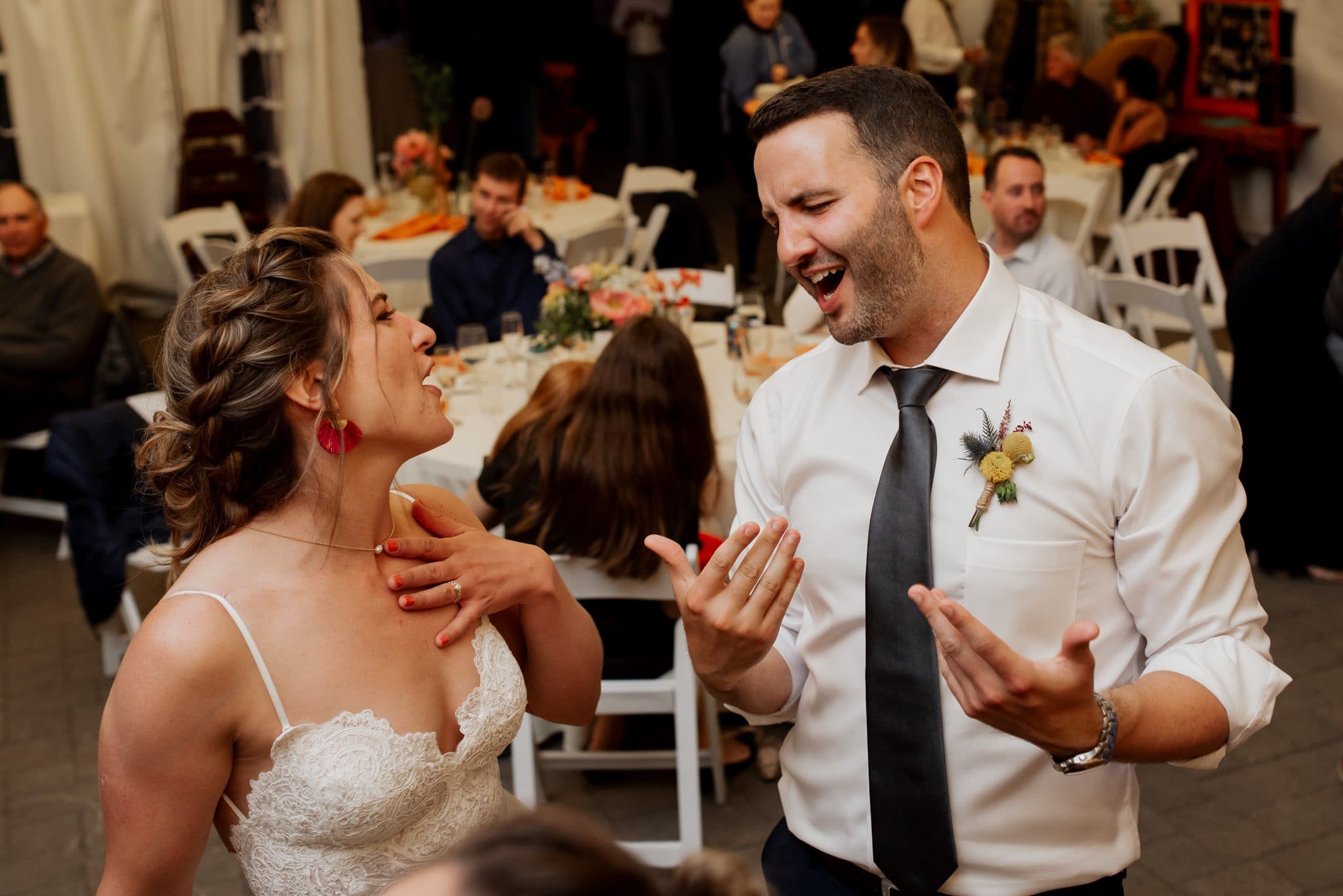 A joyful bride in a white lace dress and a man in a crisp white shirt and black tie laugh and sing at their picturesque Skibowl wedding reception. Surrounded by seated guests, the tables are elegantly decorated with vibrant flowers, capturing the essence of this enchanting celebration.