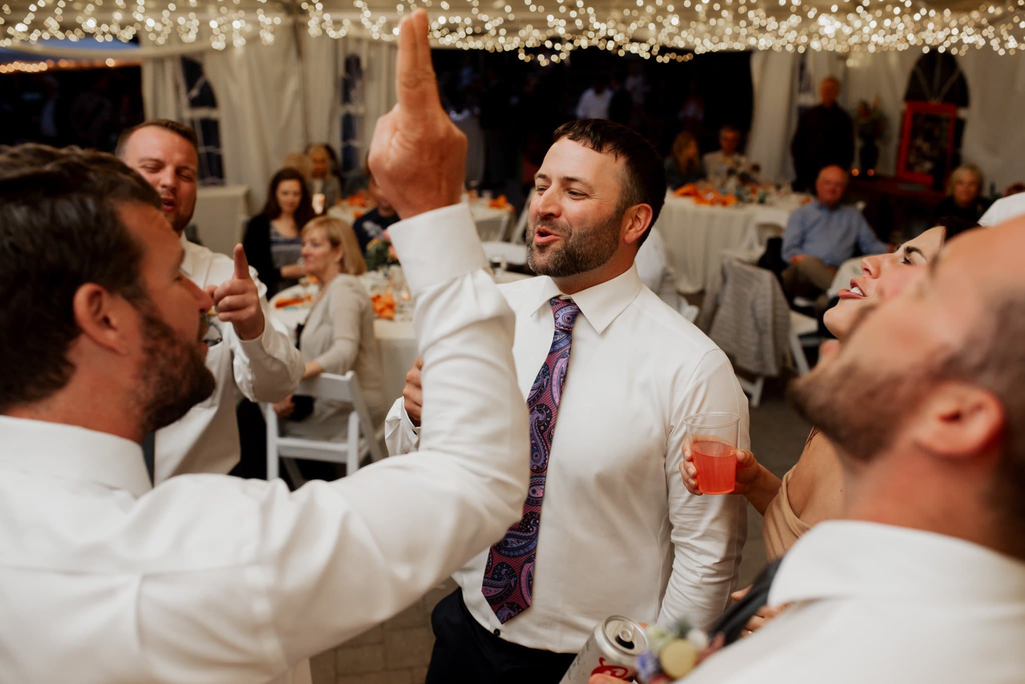 A group of people dressed in formal attire are cheerfully singing or talking, some holding drinks. They are inside a tent adorned with string lights, capturing the joyous essence of Skibowl wedding photos. Tables with seated guests gleefully enjoying the celebration are visible in the background.