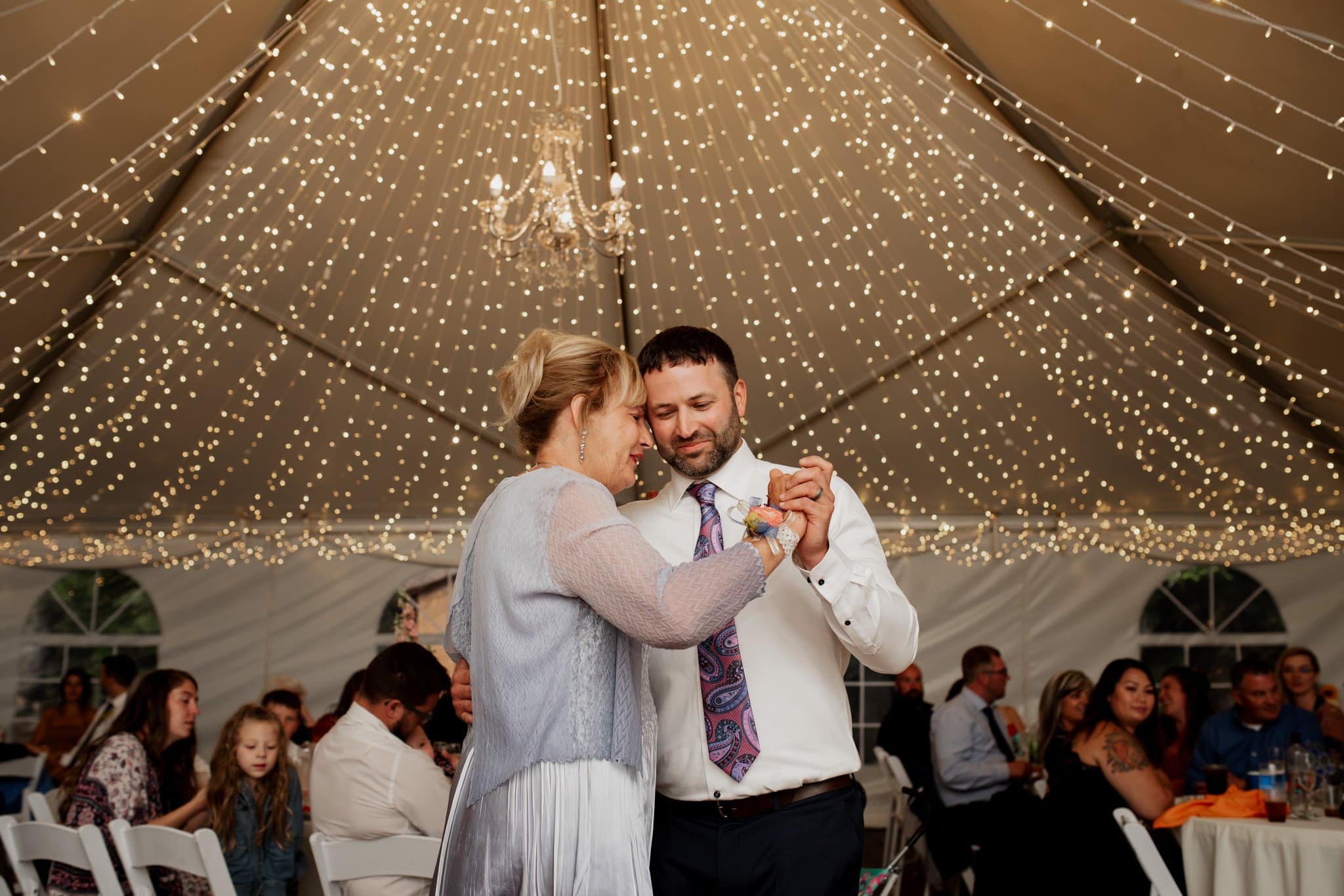 A couple dances under a canopy of twinkling string lights in a tent at their Mt Hood Ski Bowl wedding. The man, wearing a shirt and tie, holds the woman in a light dress as seated guests enjoy the festive ambiance in the background.
