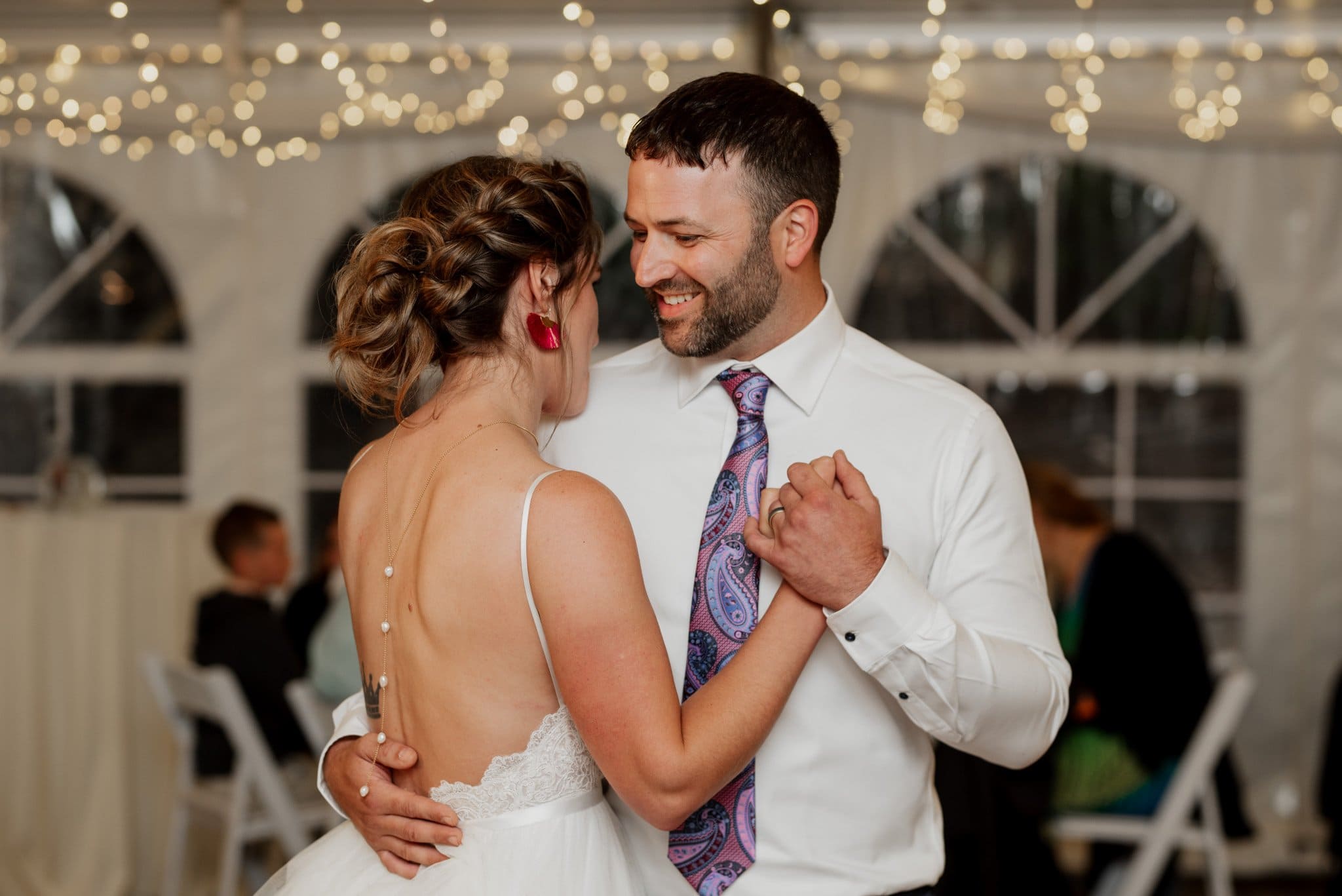 At a Mt. Hood Ski Bowl wedding, a couple dances gracefully at the reception. The bride stuns in a backless white dress, while the groom sports a white shirt with a colorful tie. String lights twinkle above them, creating a magical atmosphere as guests are seated in the background.