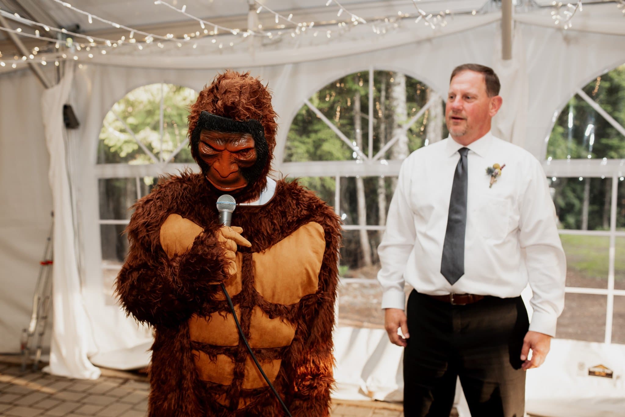 Inside a tent adorned with string lights, a person in a gorilla costume holds a microphone before the seated guests at a Mt Hood Ski Bowl wedding. Beside them stands a man in a white shirt and black tie, smiling warmly as trees sway gently outside.