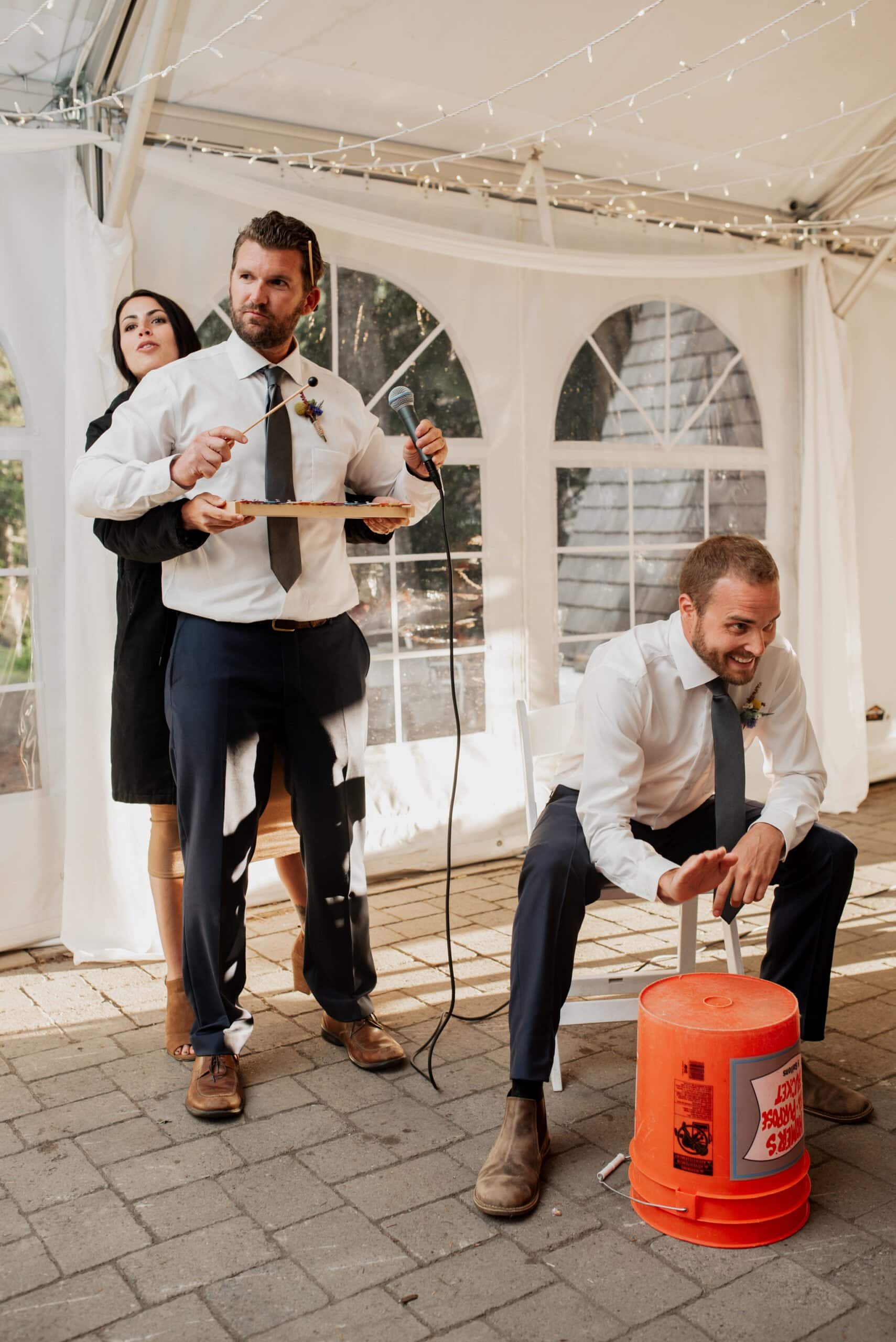 Three people are indoors under a tent at a Mt Hood Ski Bowl wedding. Two men stand while one sits on an orange bucket. One man holds a microphone and a tray, assisted by a woman behind him. The setting features elegant white drapes and arched windows, adding charm to the celebration.