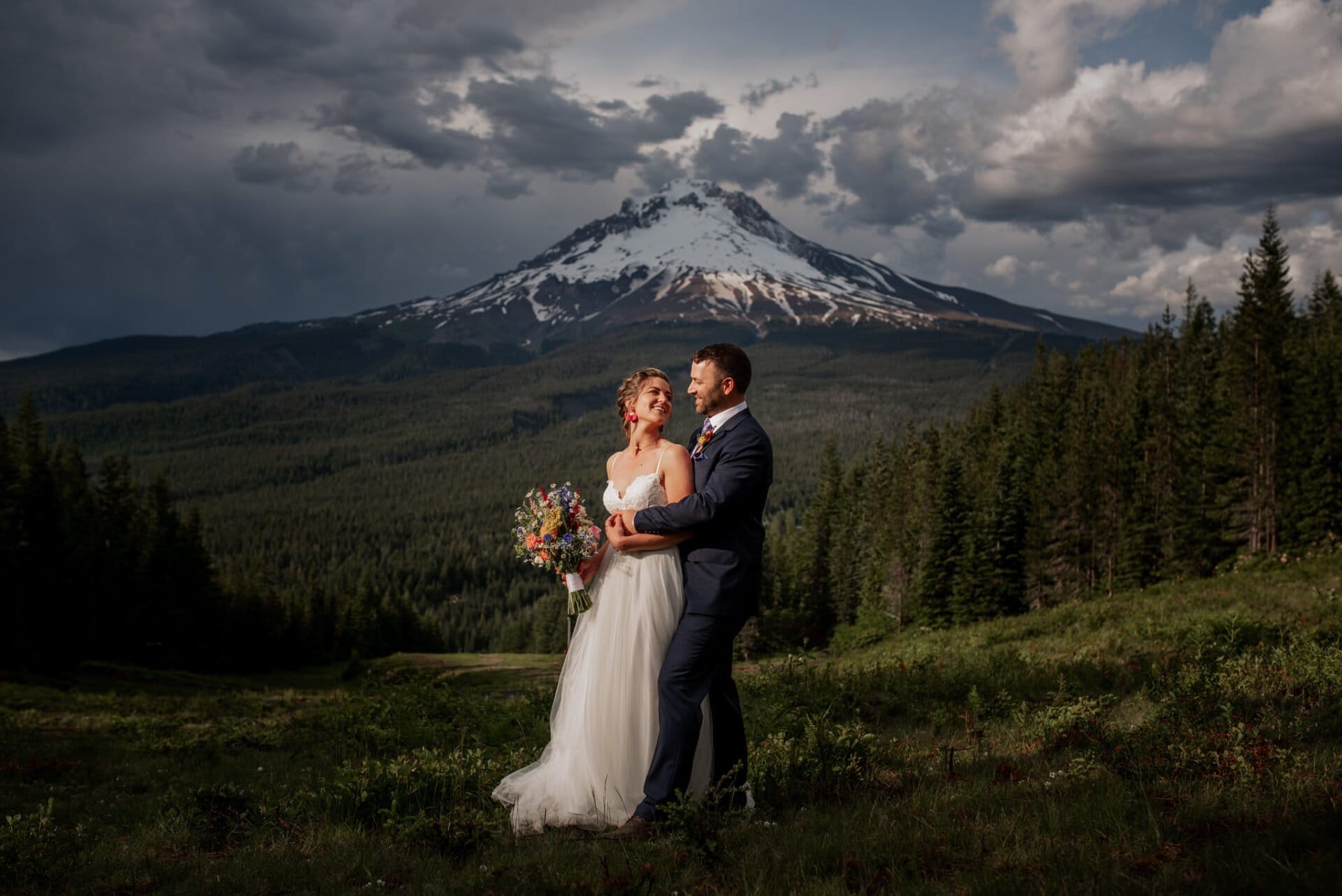 A bride and groom stand joyfully in a lush green field at Mt. Hood Ski Bowl wedding, the bride clutching her bouquet. Behind them, a dramatic sky frames the snow-capped peaks of Mt. Hood, while a dense forest envelops the serene scene.
