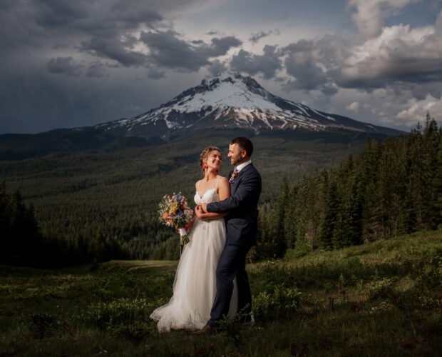 A bride and groom stand joyfully in a lush green field at Mt. Hood Ski Bowl wedding, the bride clutching her bouquet. Behind them, a dramatic sky frames the snow-capped peaks of Mt. Hood, while a dense forest envelops the serene scene.