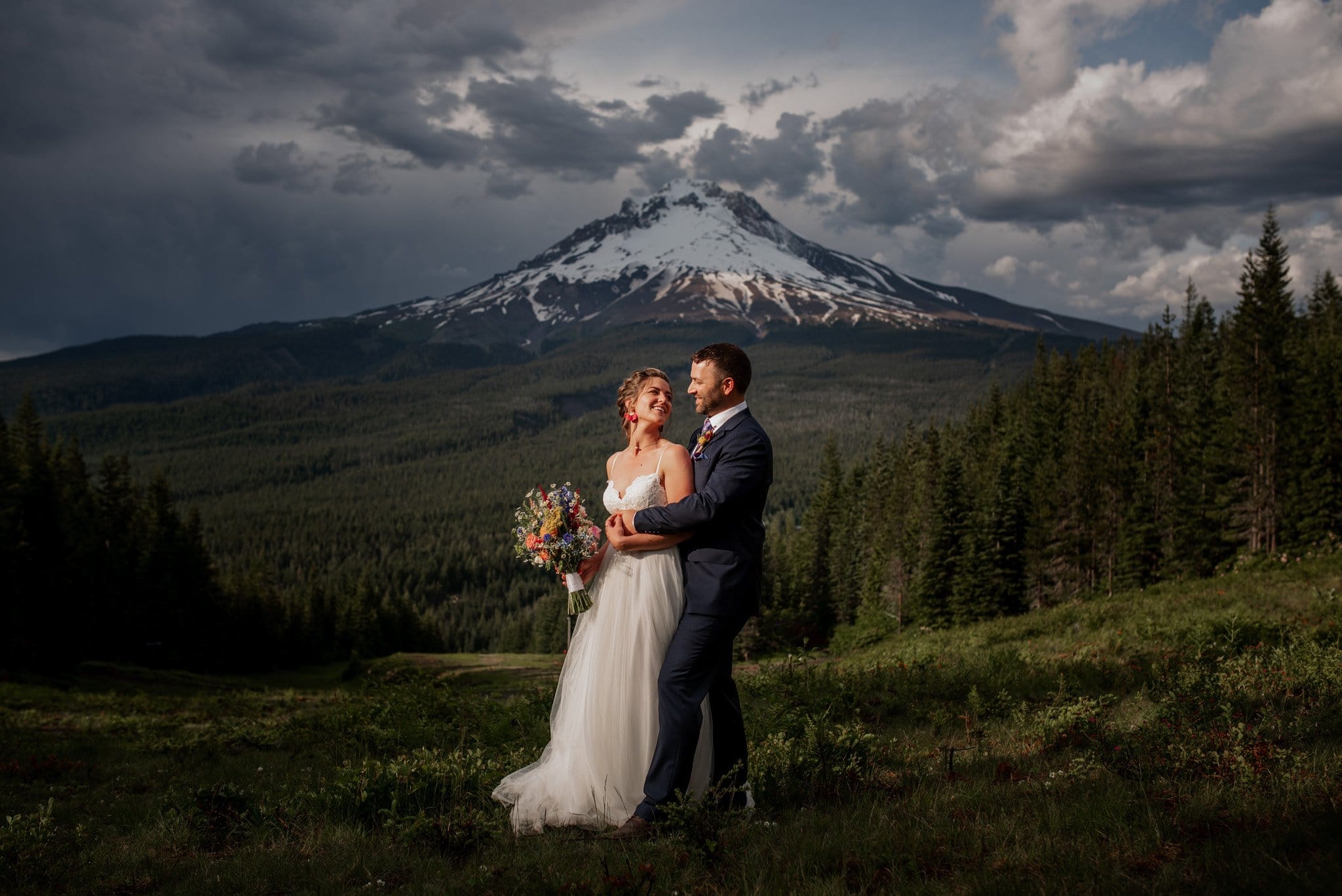 A bride and groom stand joyfully in a lush green field at Mt. Hood Ski Bowl wedding, the bride clutching her bouquet. Behind them, a dramatic sky frames the snow-capped peaks of Mt. Hood, while a dense forest envelops the serene scene.