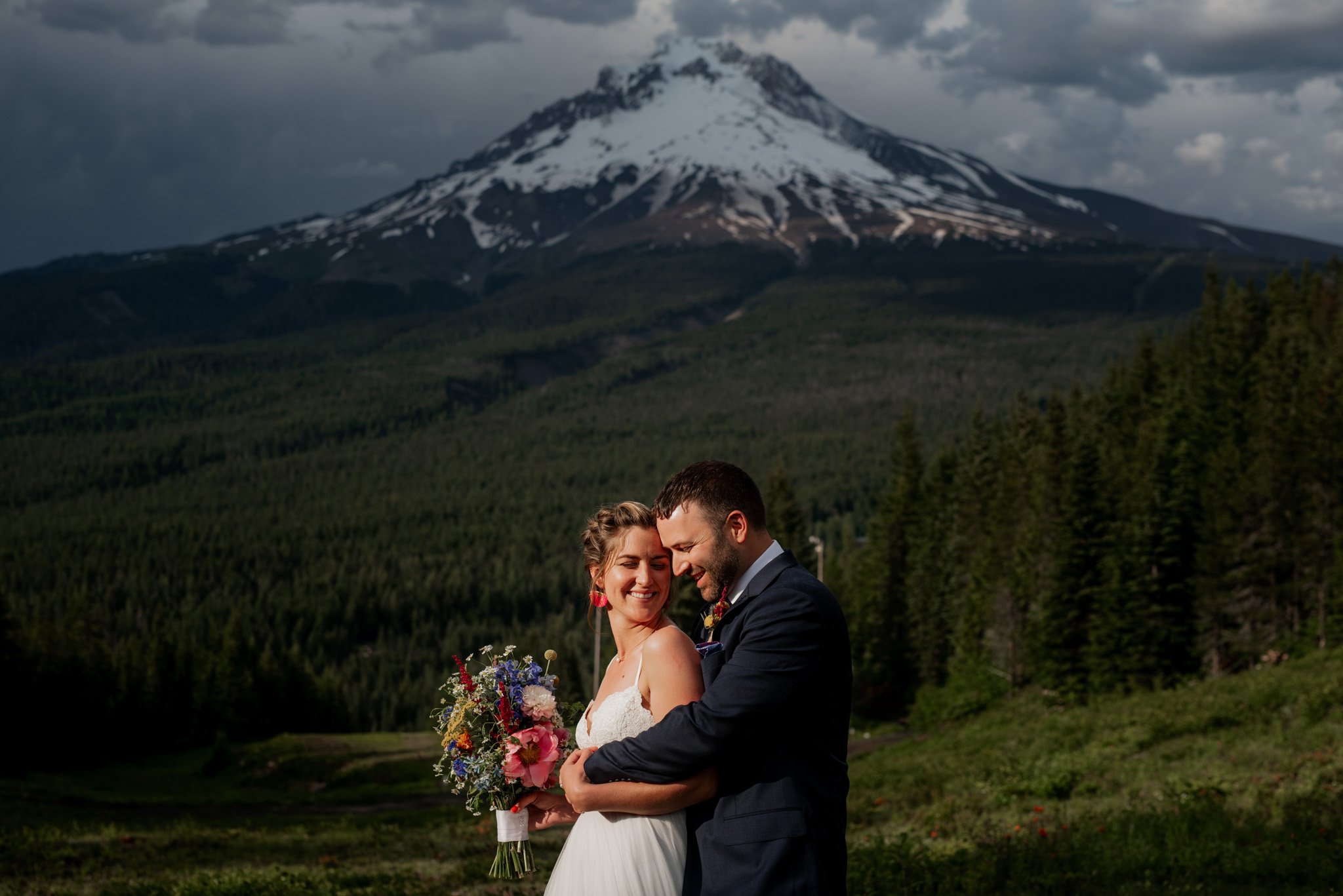 A couple embraces outdoors with Mt. Hoods majestic backdrop. The bride, holding a bouquet of colorful flowers, smiles while the groom stands behind her, looking content. Snow partly covers the mountainside near Ski Bowl and is surrounded by a lush green forest.