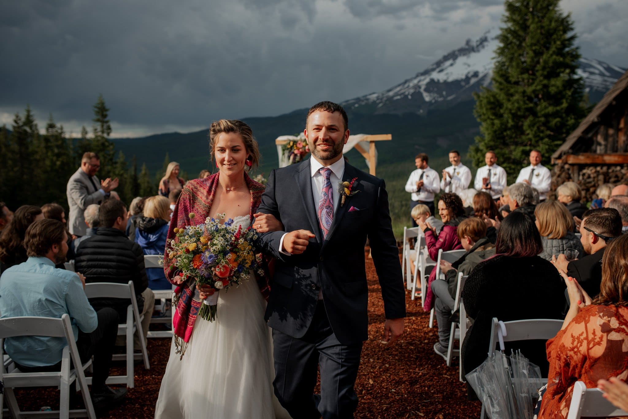 The bride and groom smile as they walk down the aisle of their Mt. Hood Ski Bowl wedding. The bride holds a colorful bouquet and wears a white dress with a patterned shawl. Guests sit on white chairs, with a majestic snow-capped mountain under a cloudy sky in the background.