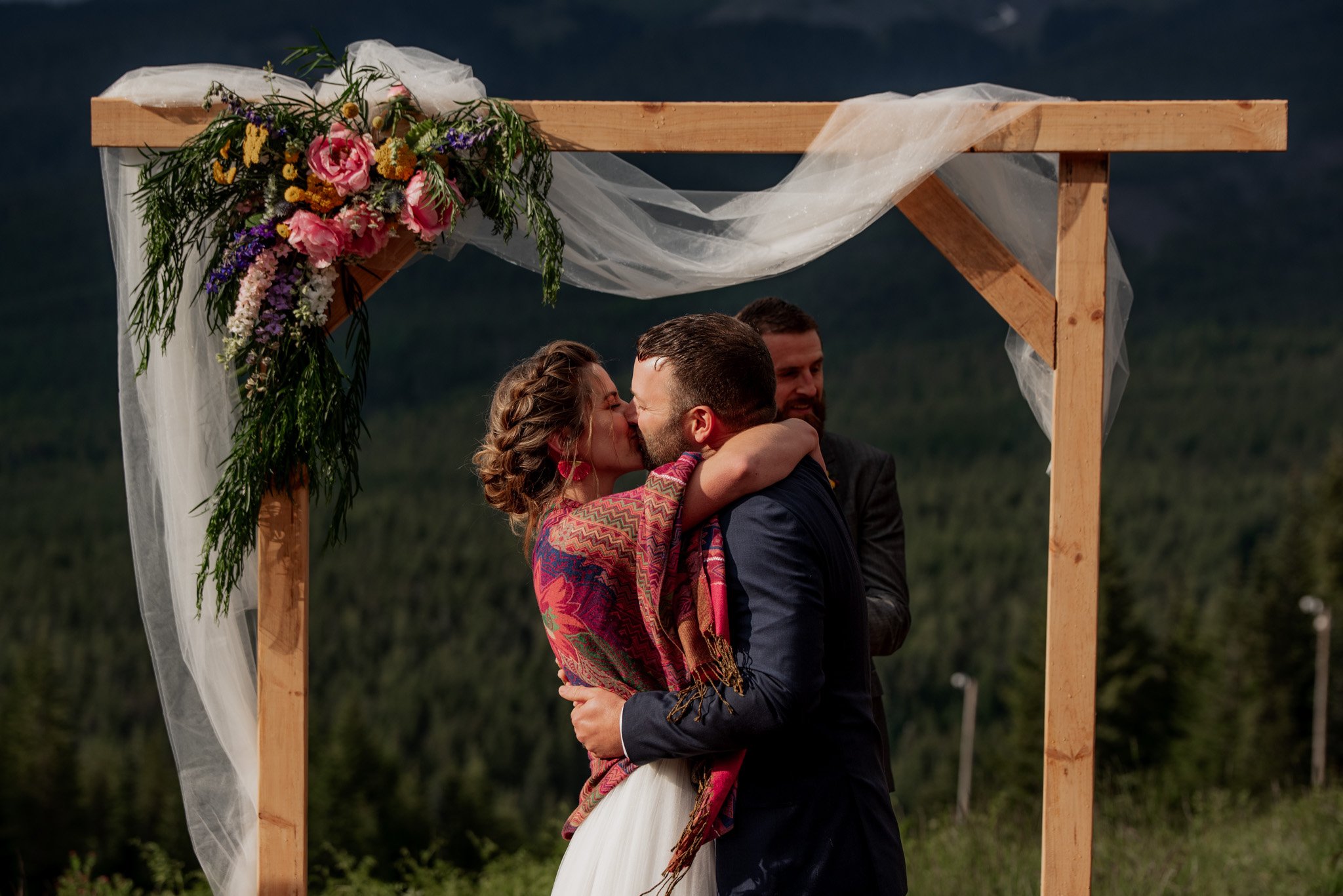 A couple kisses under a wooden arch adorned with flowers and sheer fabric at their Mt Hood Ski Bowl wedding, standing against a backdrop of lush green trees.