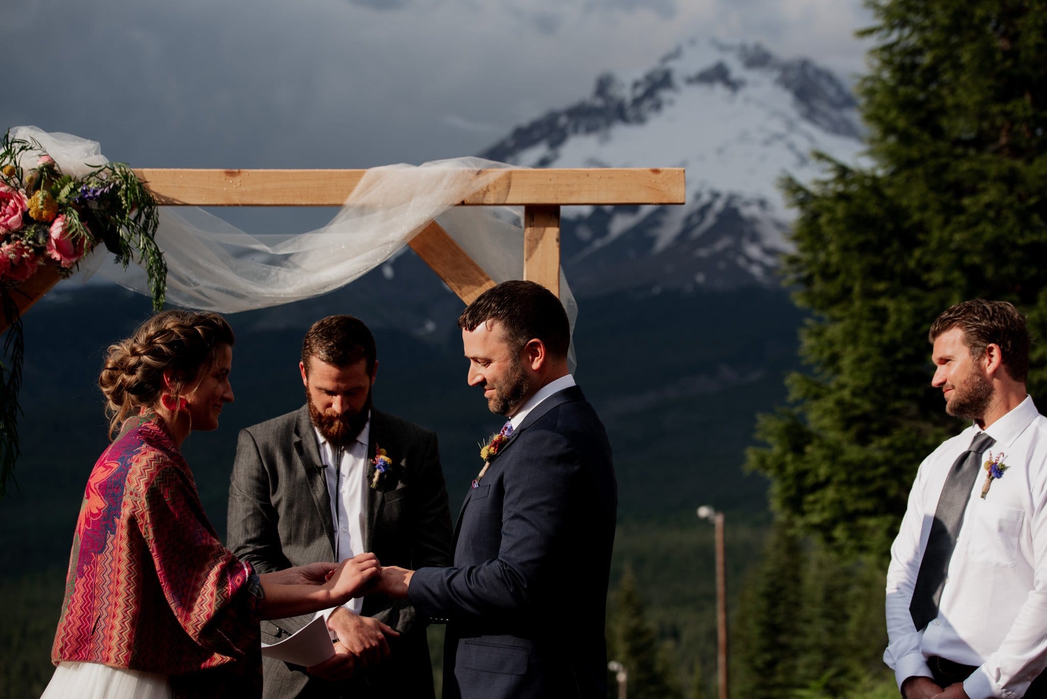 A couple exchanges rings during their outdoor Mt Hood Ski Bowl wedding. Snow-capped mountains and trees frame the moment beautifully. The groom wears a suit, while the bride drapes a colorful shawl. Two groomsmen stand nearby, dressed in white shirts and ties, completing the picturesque scene.